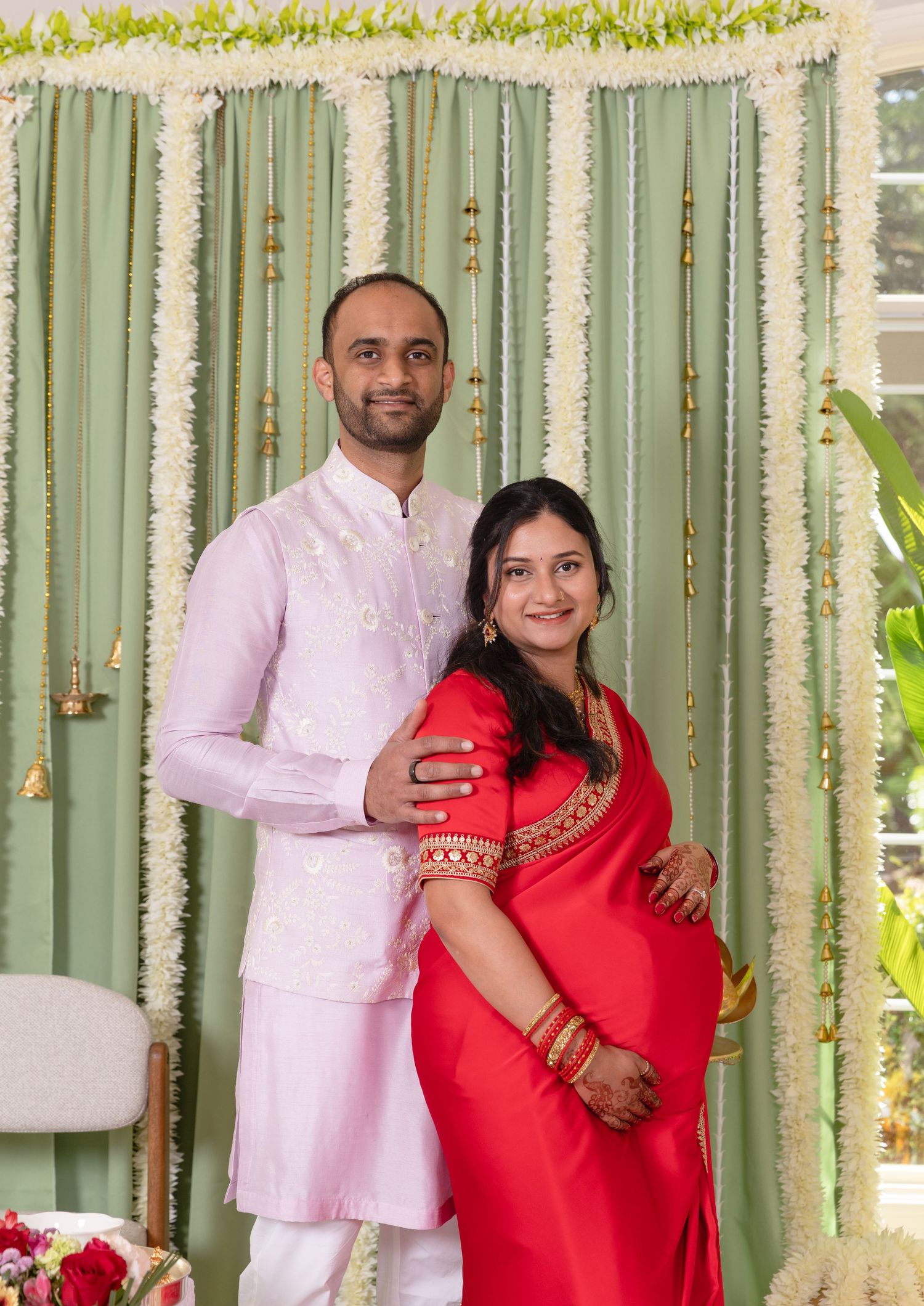 Pregnant woman in red saree with husband in pink kurta posing at traditional Indian ceremony with floral backdrop.