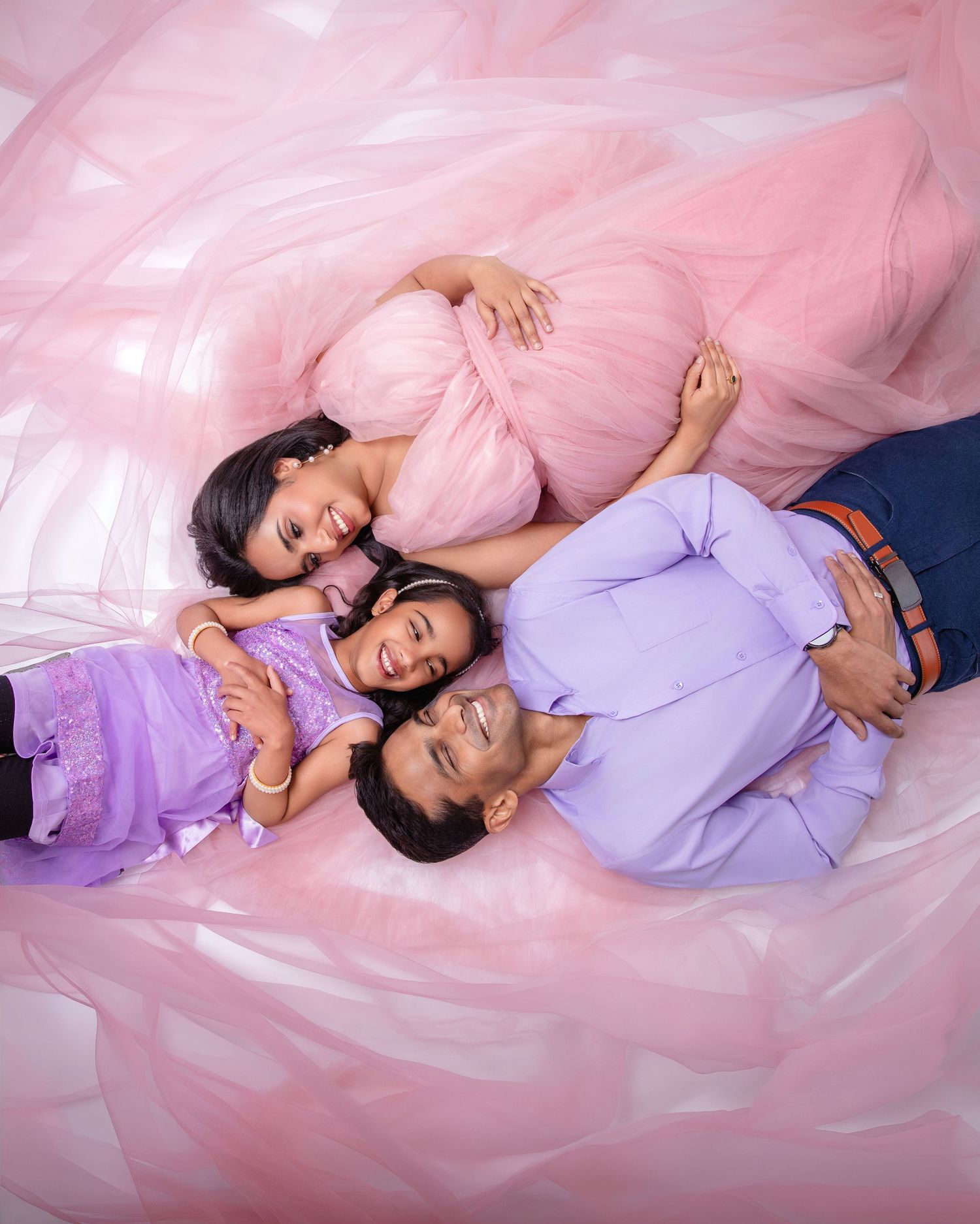 A joyful family of three lying on pink tulle fabric, with a pregnant mother in a pink gown, father, and child smiling.