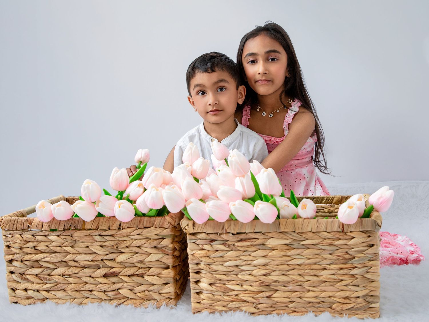Two children pose behind wicker baskets filled with pink tulips against a white background.