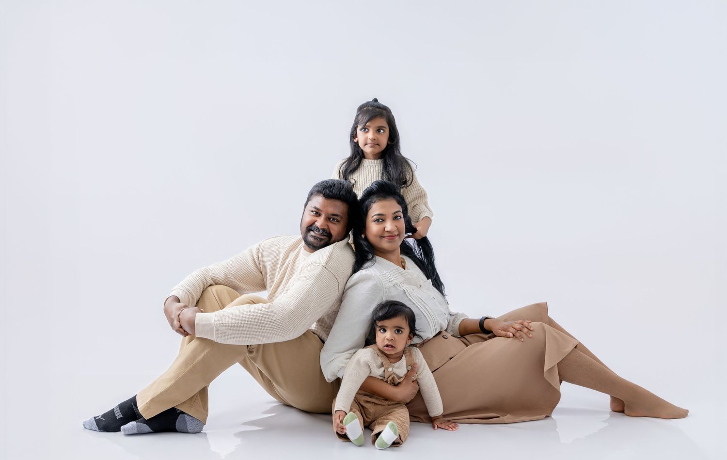 A joyful South Asian family of four poses together in a studio, wearing coordinating beige outfits on a white background.