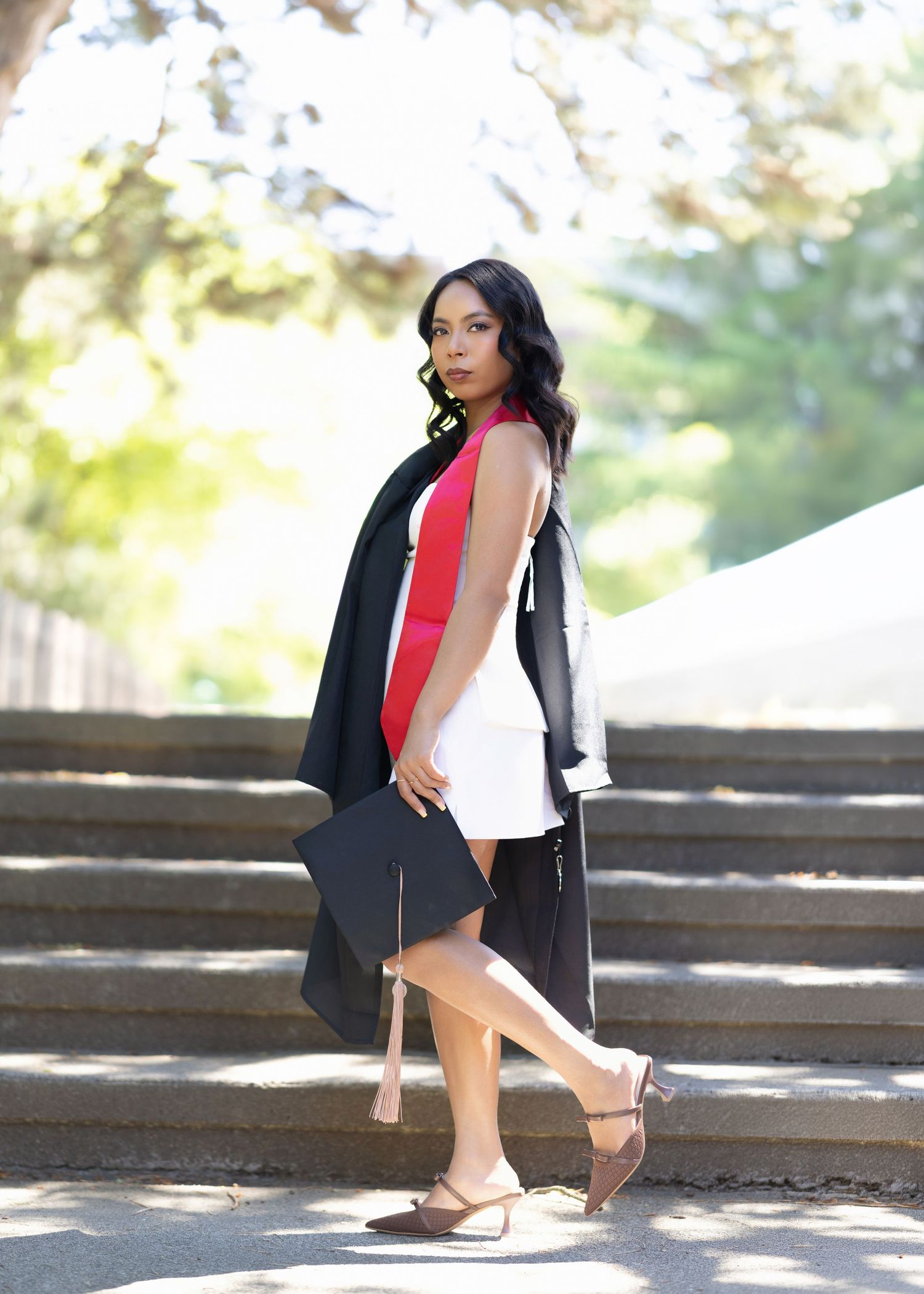 Young woman in white dress and graduation gown holds cap on outdoor steps.