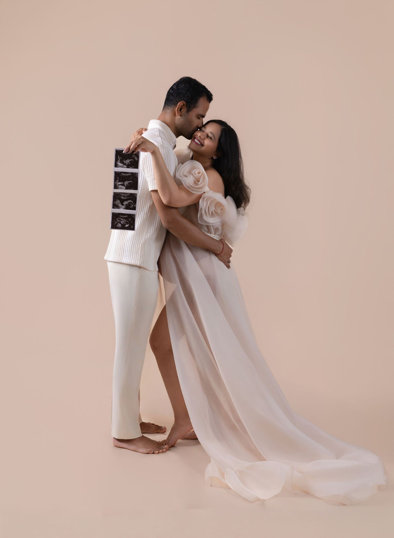 Couple in white outfits embracing during maternity photo shoot, woman in sheer gown holding ultrasound images on beige background.
