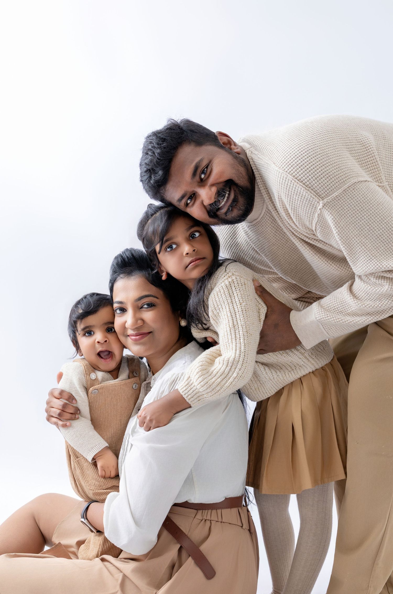 A joyful South Asian family of four poses together in coordinating beige and white outfits against a white background.