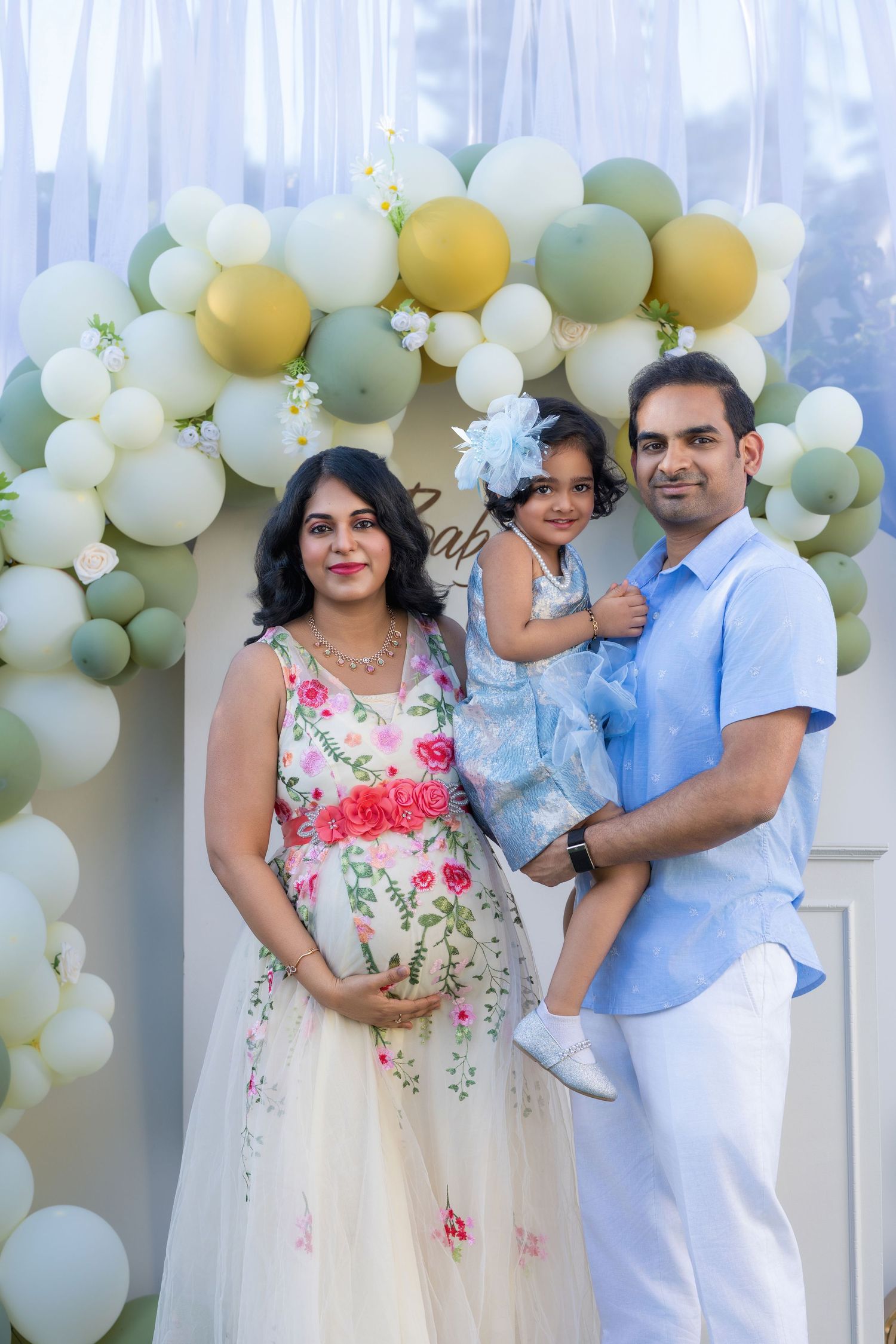 Pregnant woman in floral dress poses with husband and toddler daughter at baby shower with sage green balloon arch.