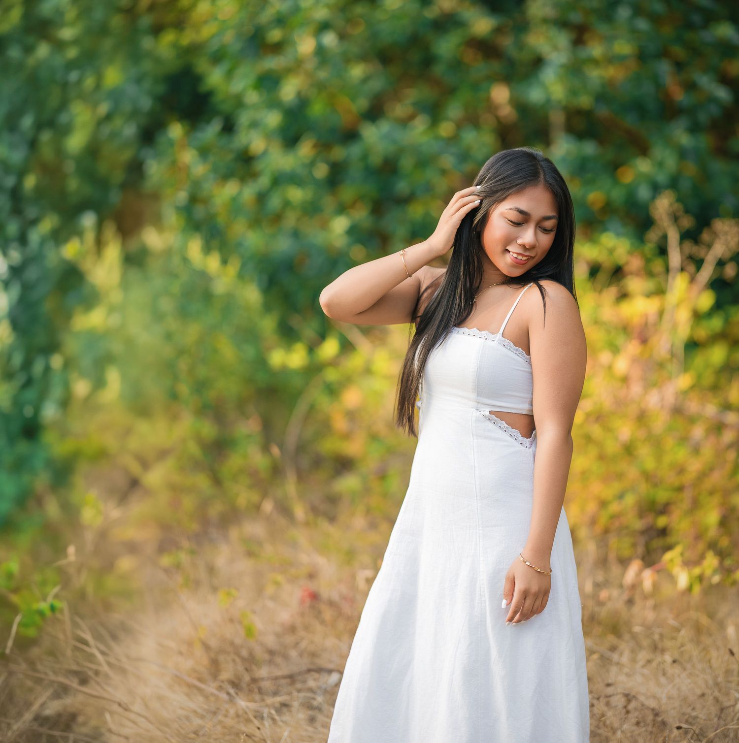 Young woman in white sundress poses gracefully outdoors among lush green trees and golden grass.