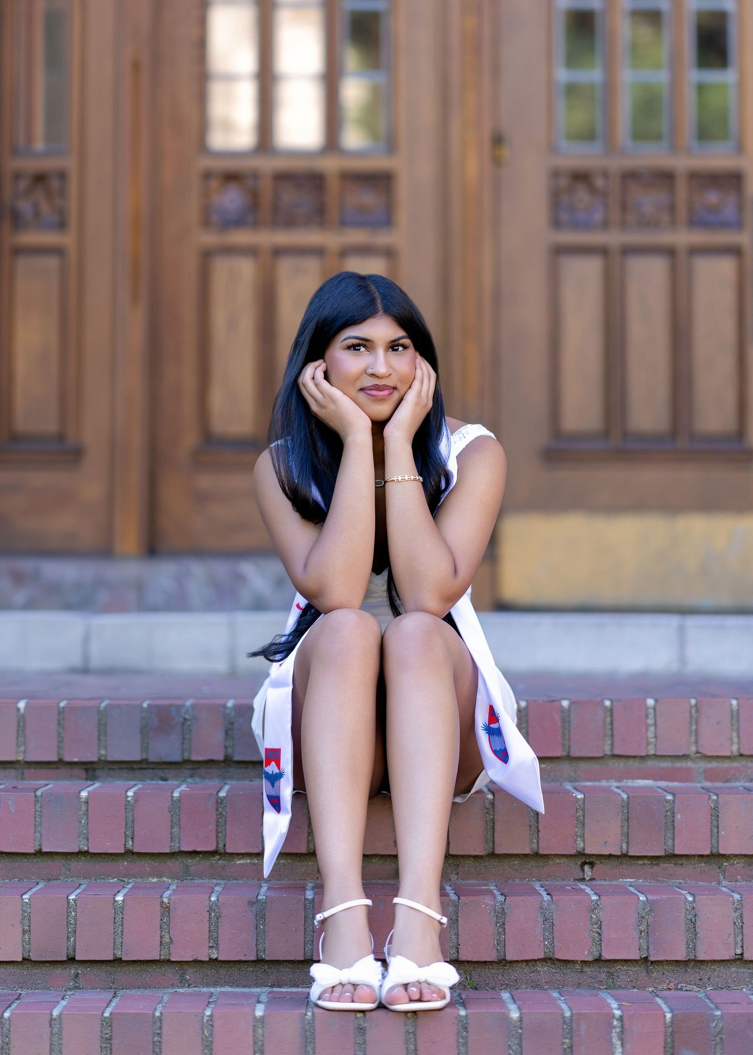 Young woman in white dress sitting on brick steps, posing thoughtfully with hands on cheeks near wooden doors.