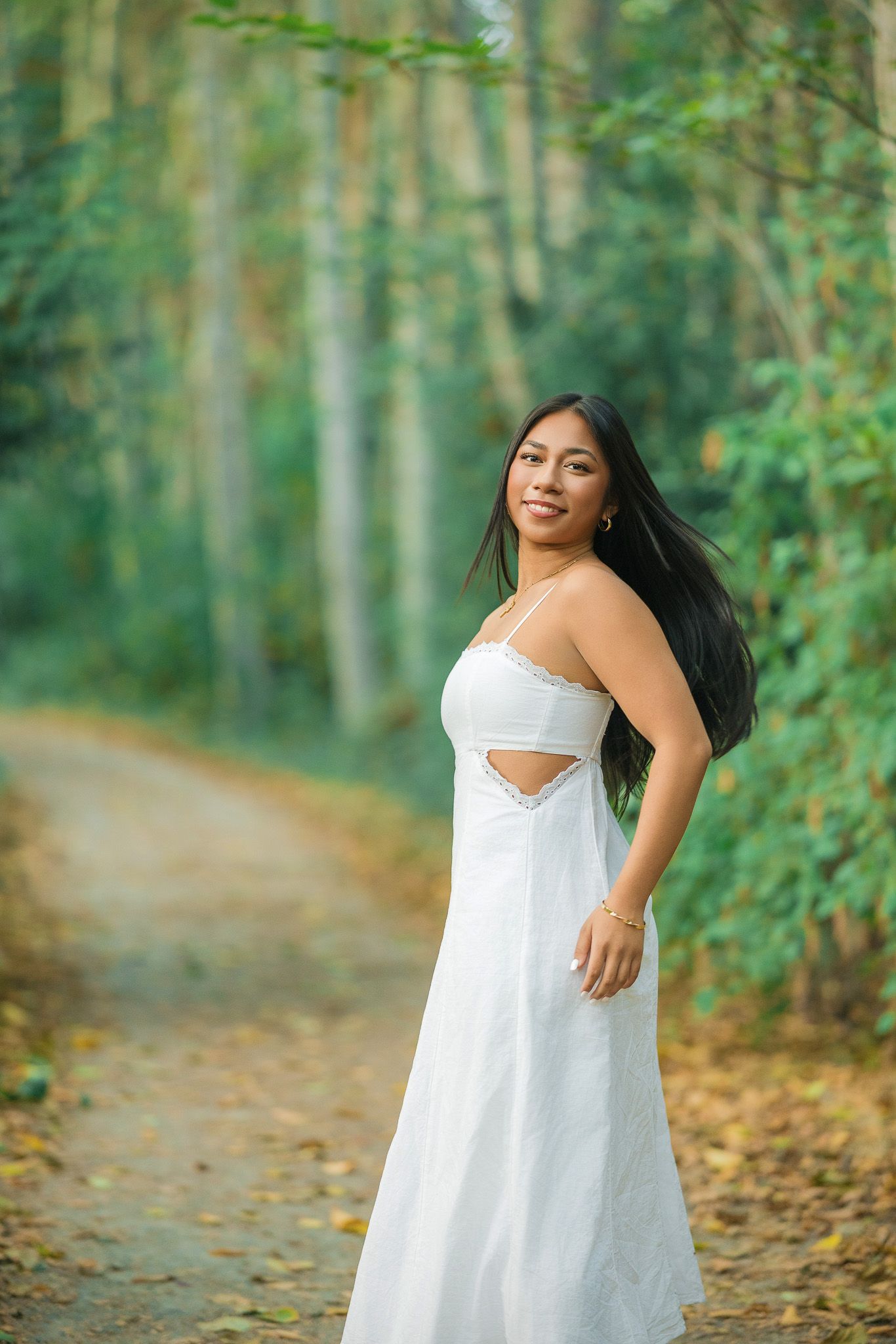 Young woman in white sundress smiling on a forest path with lush green trees in the background.