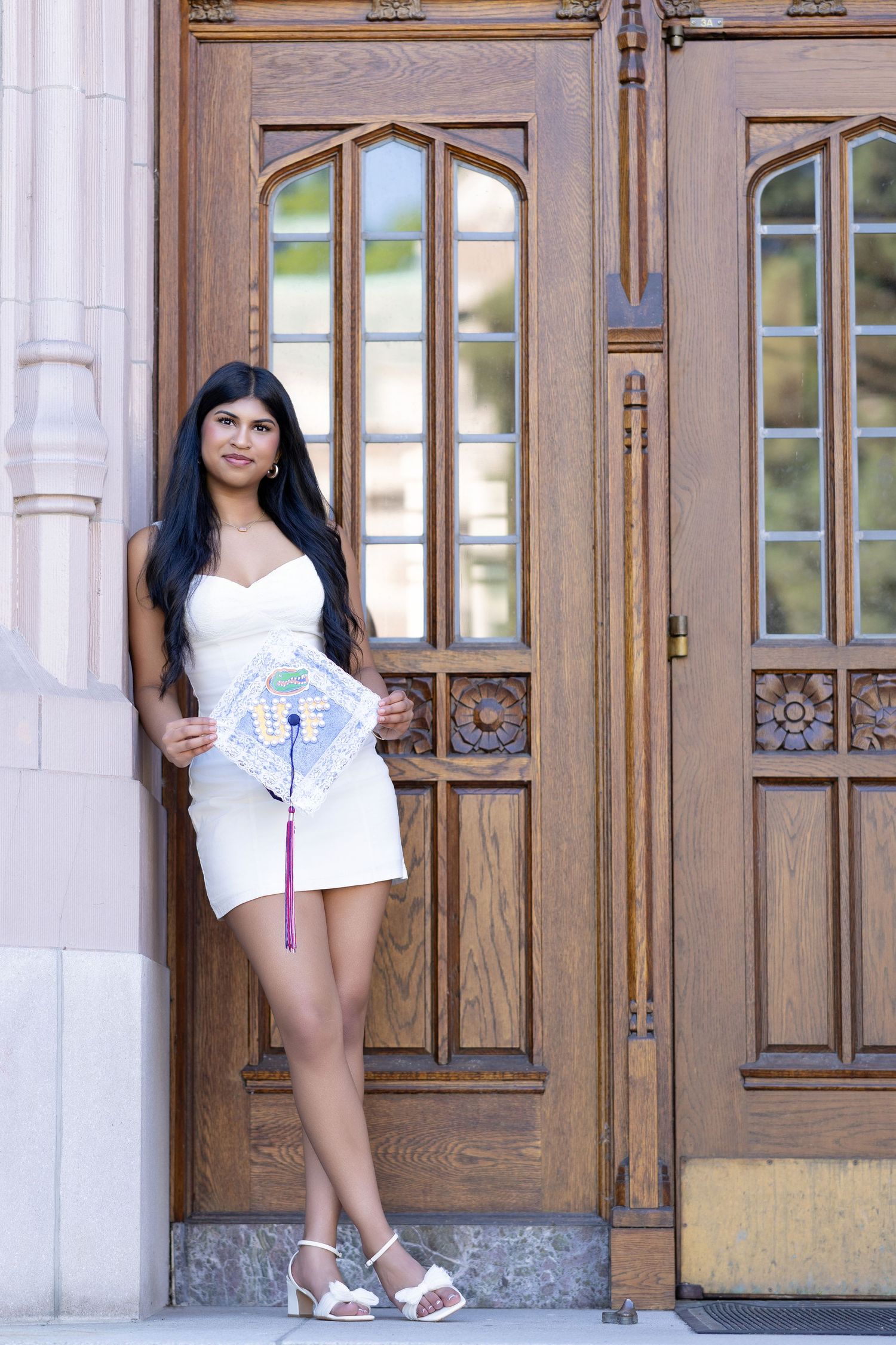 Young woman in white dress holding decorated graduation cap poses by ornate wooden doors.