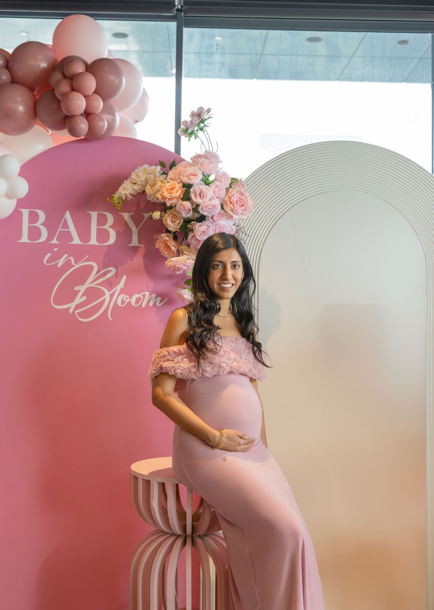 Pregnant woman in pink off-shoulder gown poses at Baby in Bloom baby shower with balloon arch and floral decor.