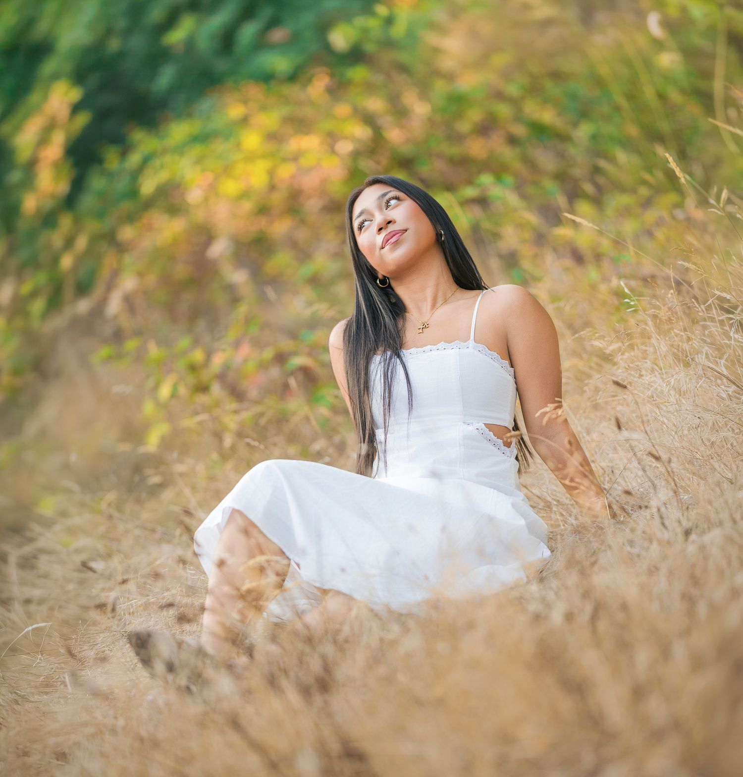 Young woman in white dress sitting in dry grass field, gazing upward with lush green foliage background.