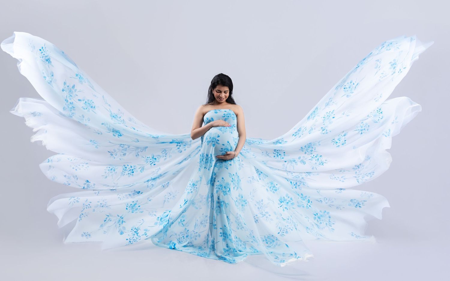 Pregnant woman in blue floral maternity gown with dramatic flowing fabric wings in studio photoshoot.