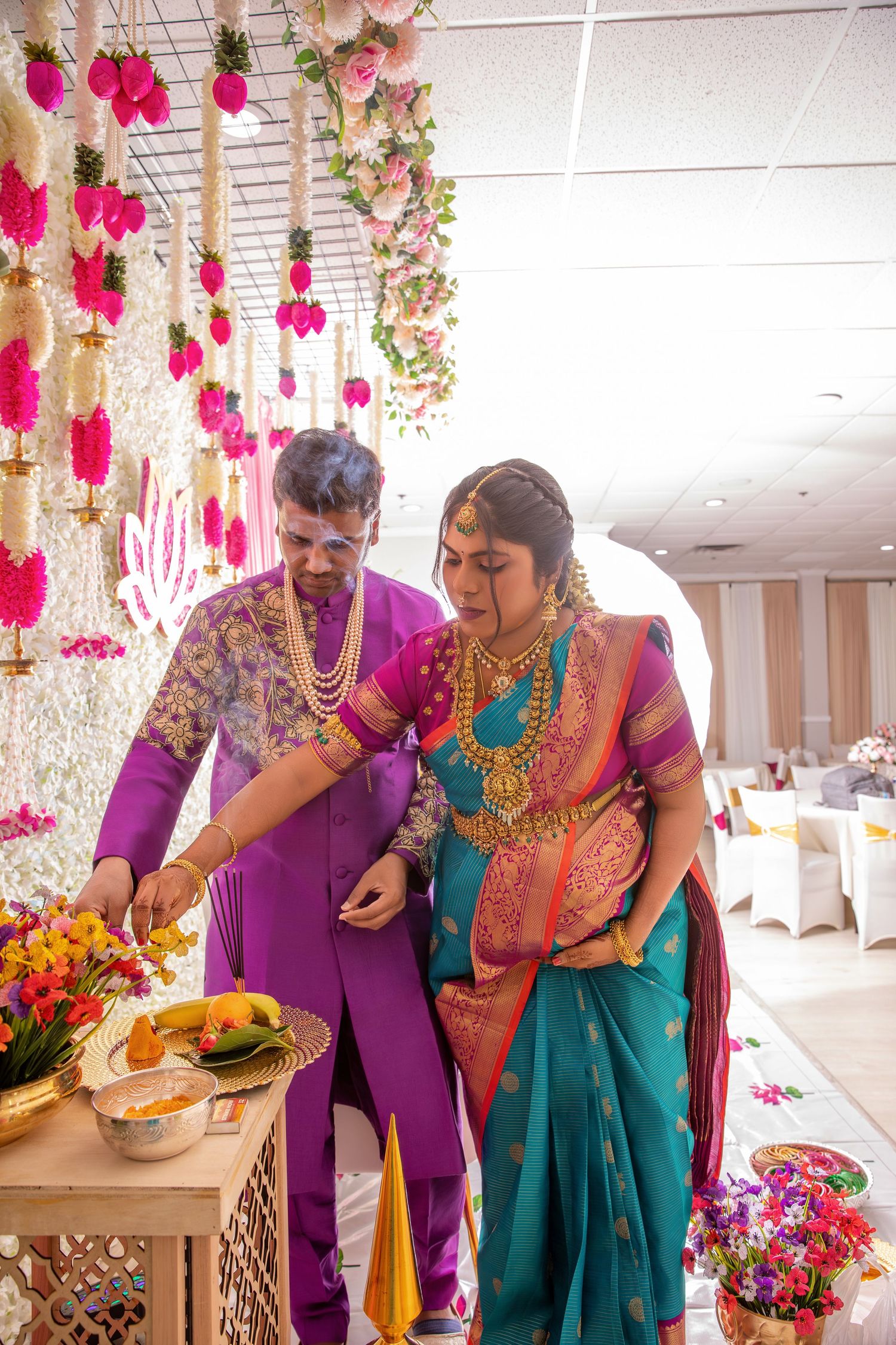 Indian bride in teal saree and groom in purple sherwani performing traditional wedding rituals under floral decorations.