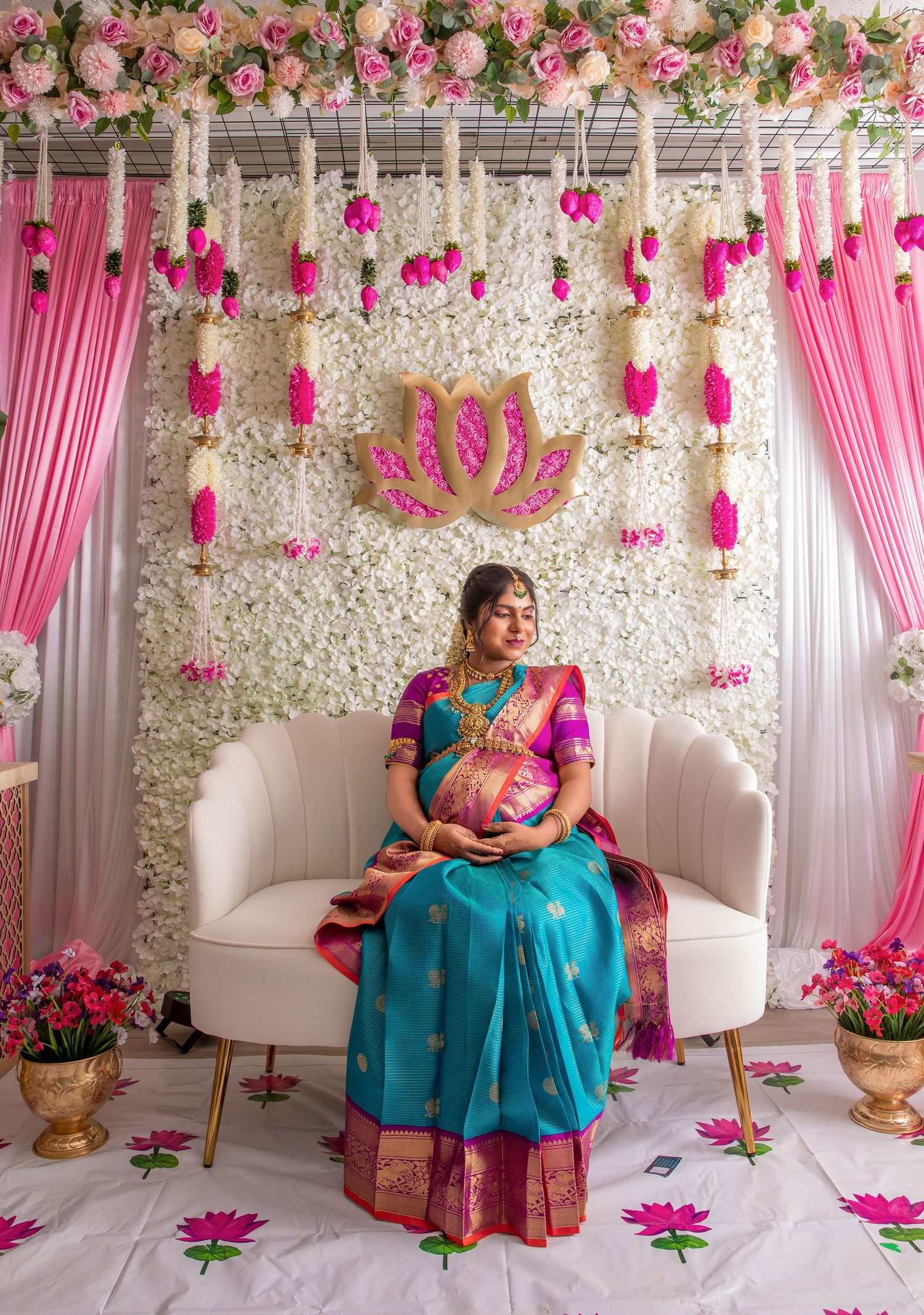 Pregnant woman in teal silk saree sits on white chair before floral backdrop with pink curtains and lotus decor.