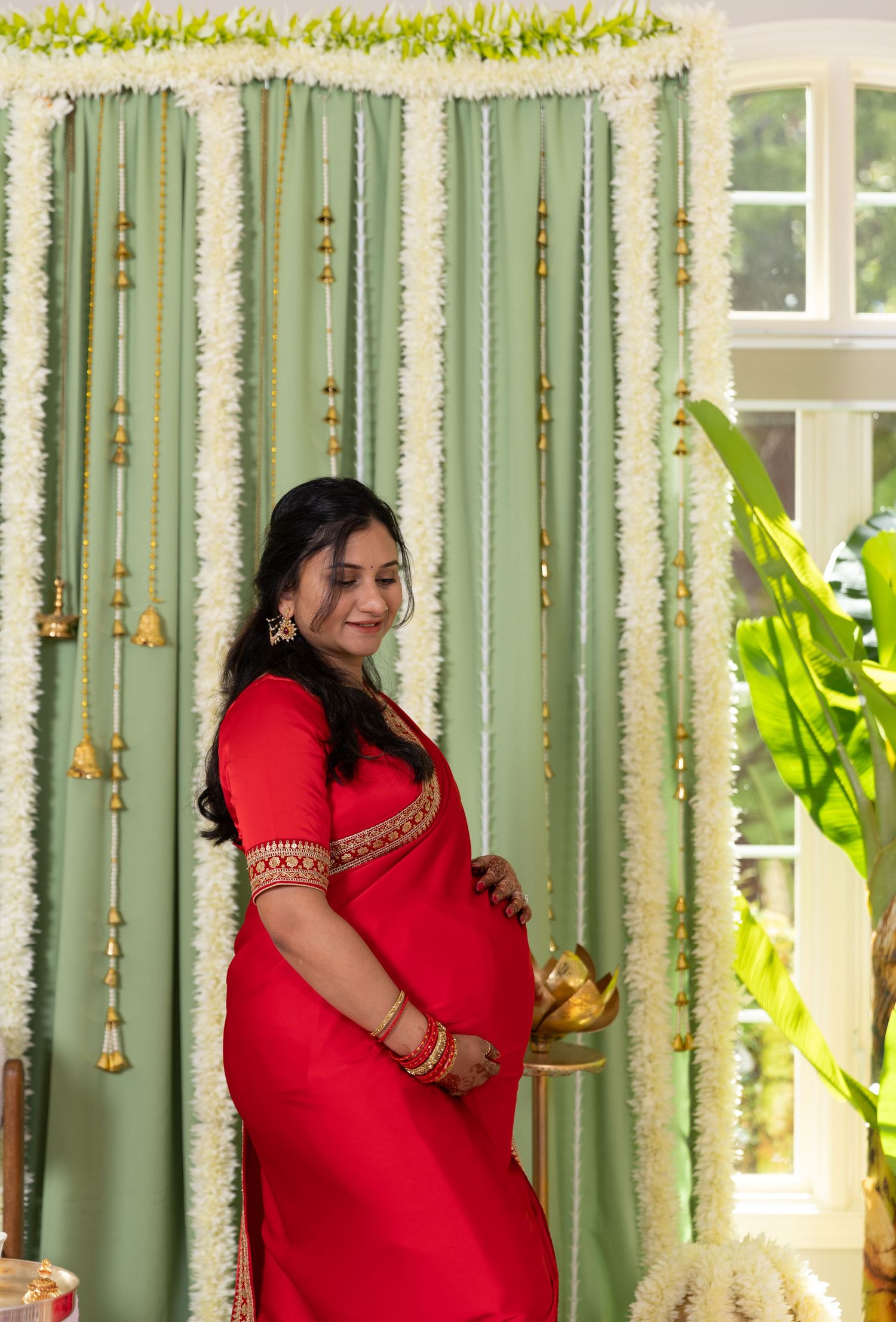 Pregnant woman in red saree holding a brass pot at a traditional Indian baby shower ceremony with floral backdrop.