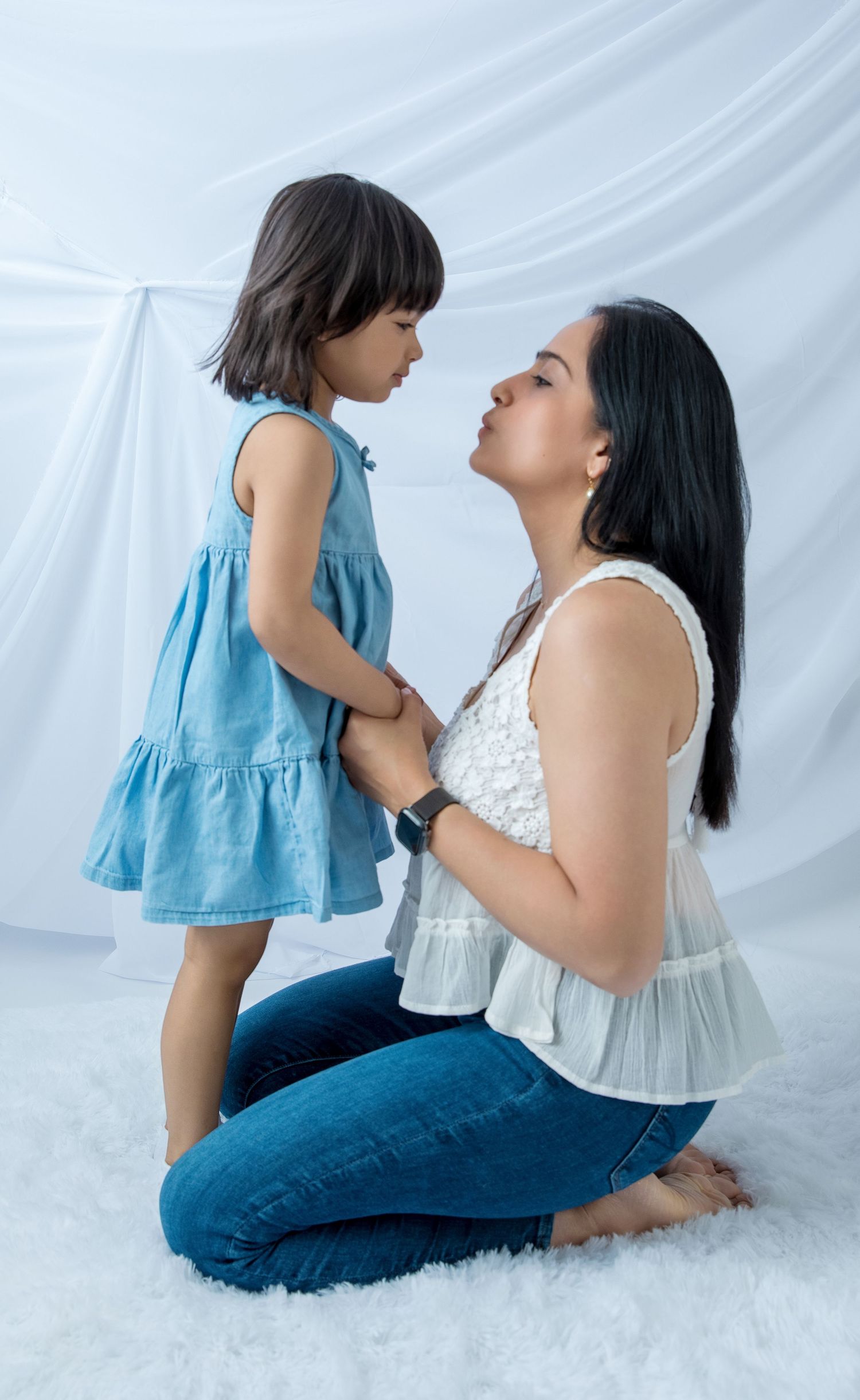 A mother kneels on a white rug, holding her young daughter's hands and leaning in for a kiss in a studio.