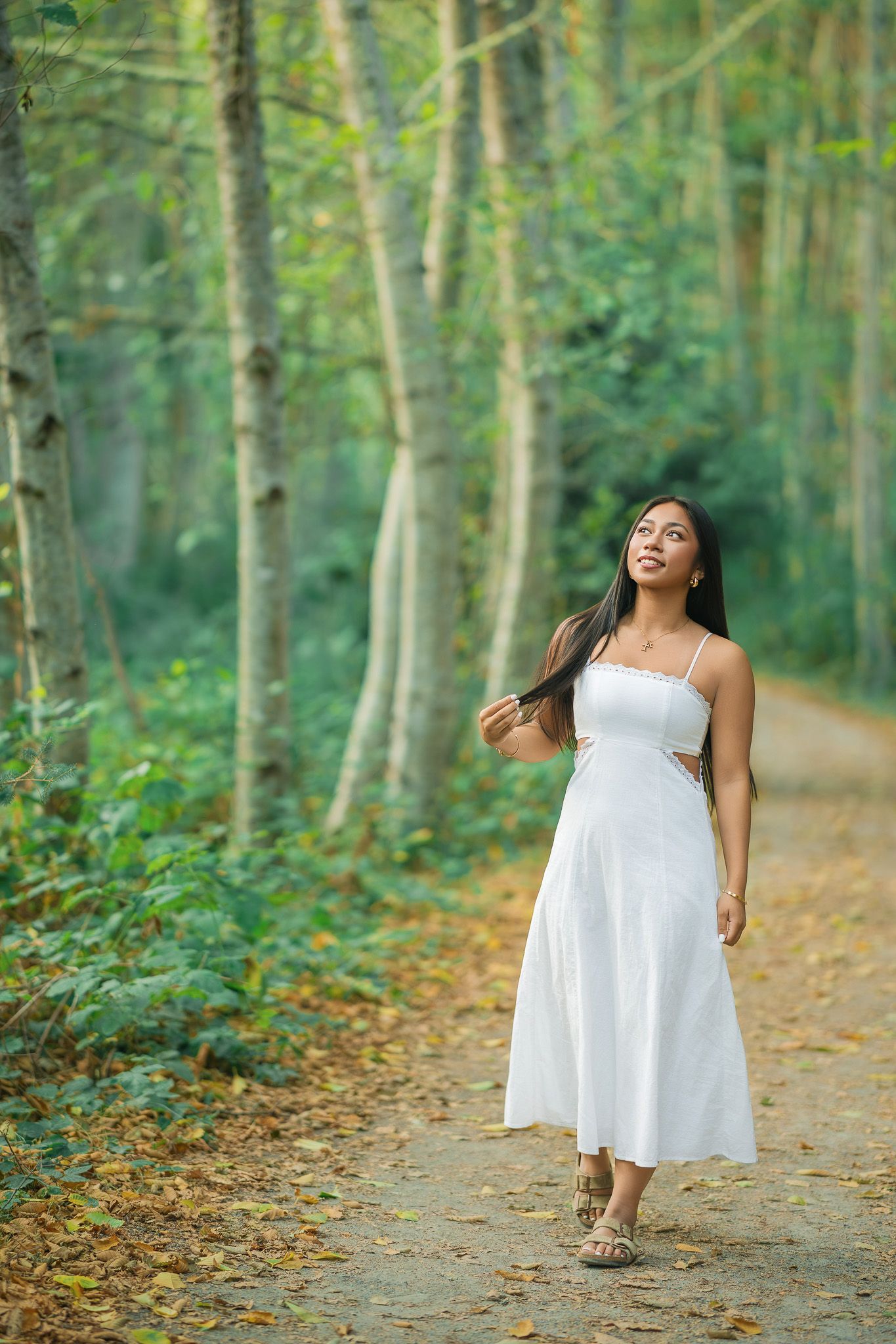 Young woman in white sundress walking on a forest path surrounded by lush green trees and autumn leaves.