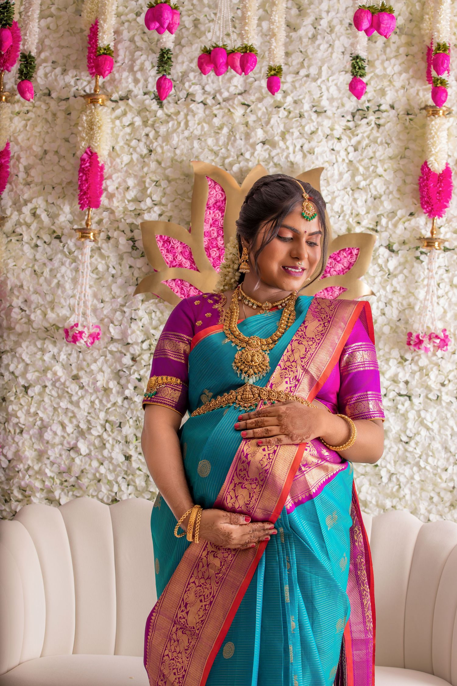 Pregnant woman in teal and pink silk saree with gold jewelry poses before floral backdrop.