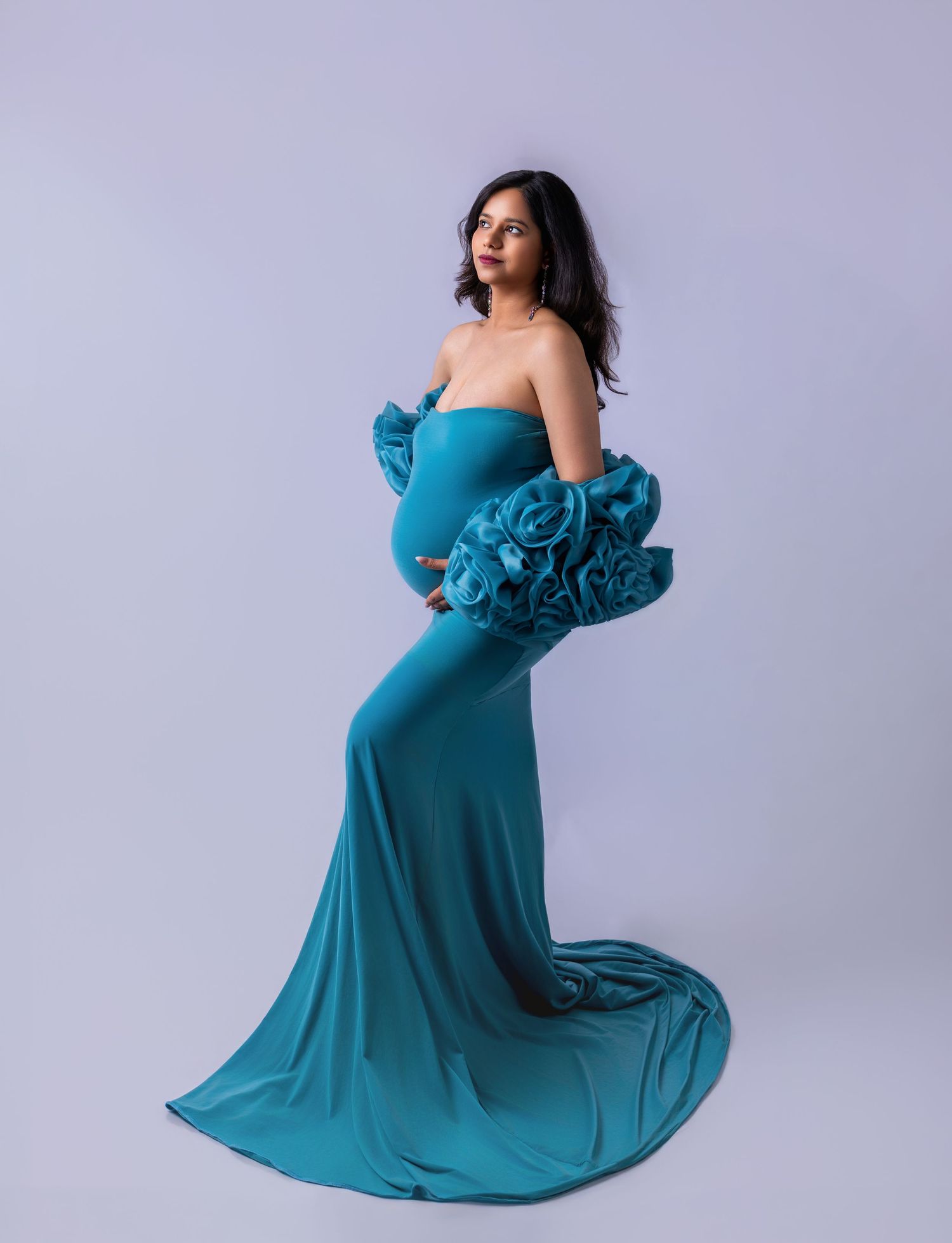 Pregnant woman in teal strapless maternity gown with floral ruffle detail, posing sideways in studio.