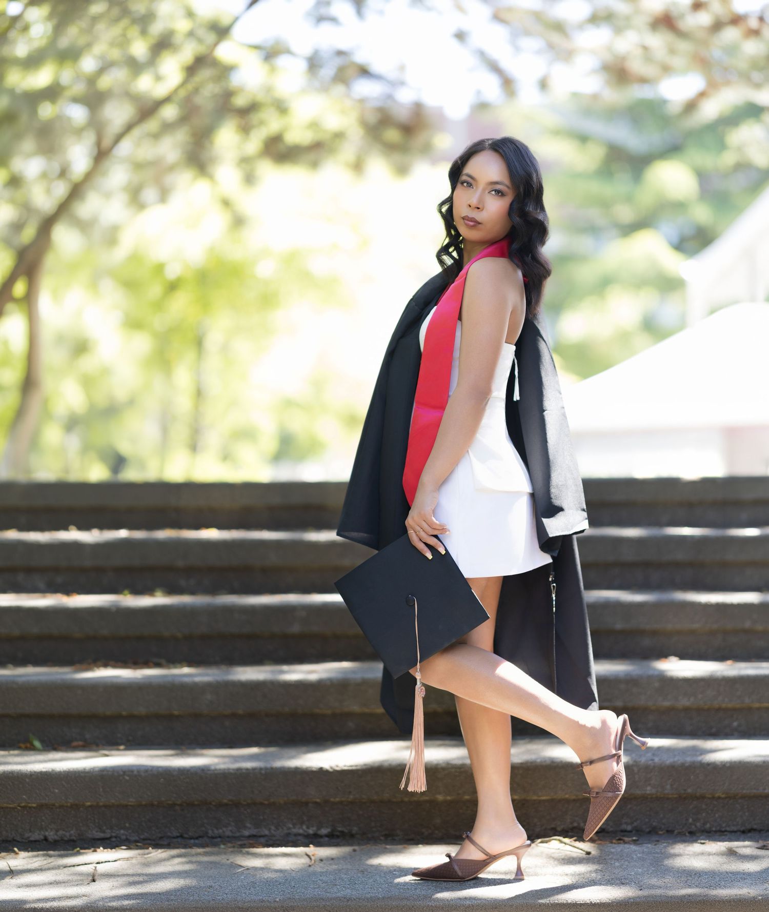 Young woman in graduation gown and white dress poses gracefully on outdoor steps surrounded by lush green trees.
