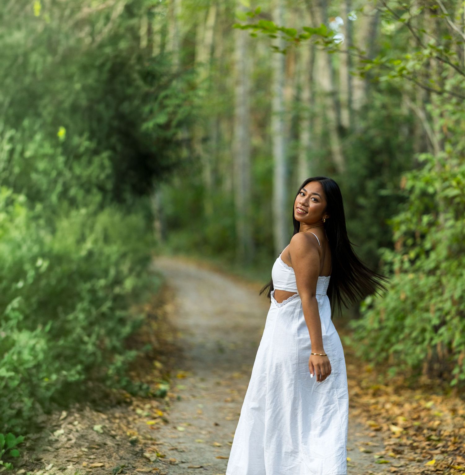 A young woman in a white maxi dress stands on a forest path surrounded by lush green trees.