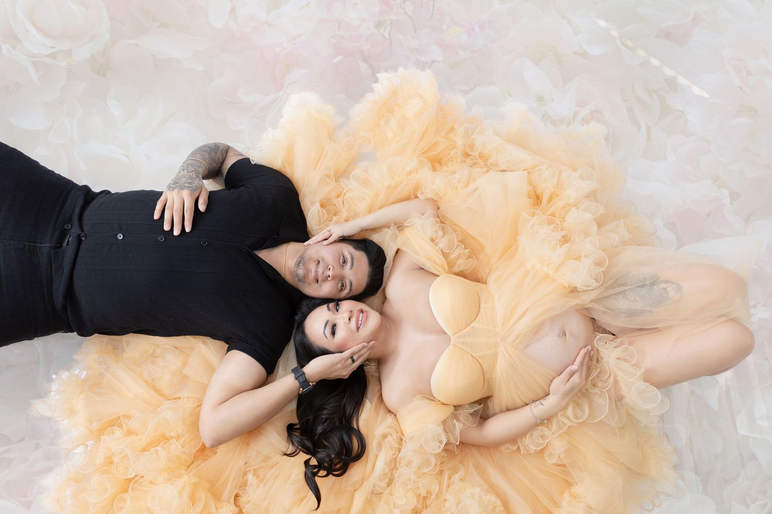 Couple lying together on floral floor, woman in golden tulle maternity gown during pregnancy photoshoot.