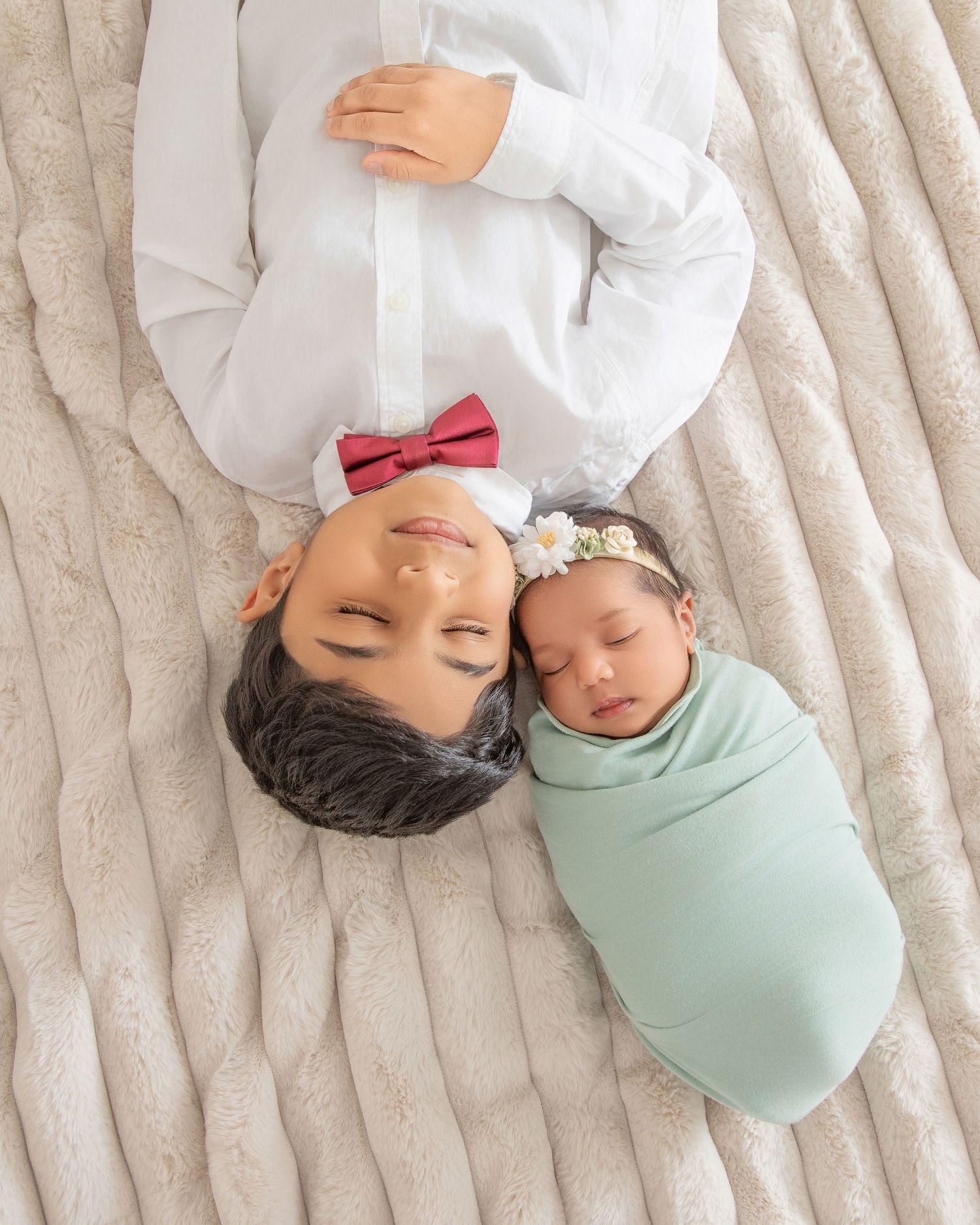 A boy in a white shirt and red bow tie lies beside a swaddled newborn baby in mint green on a soft blanket.