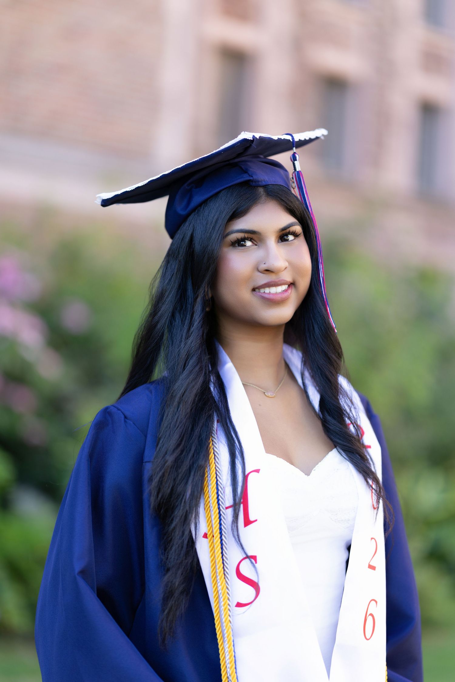Young woman in navy blue graduation cap and gown smiling outdoors, wearing honor stole with red lettering.