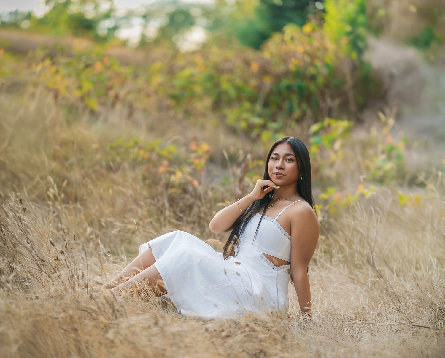 Young woman in white dress sitting in golden dry grass field with colorful foliage background.