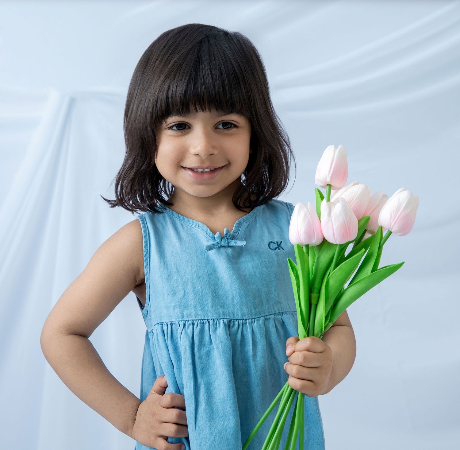 A smiling young girl in a blue Calvin Klein dress holds pink tulips against a light blue background.