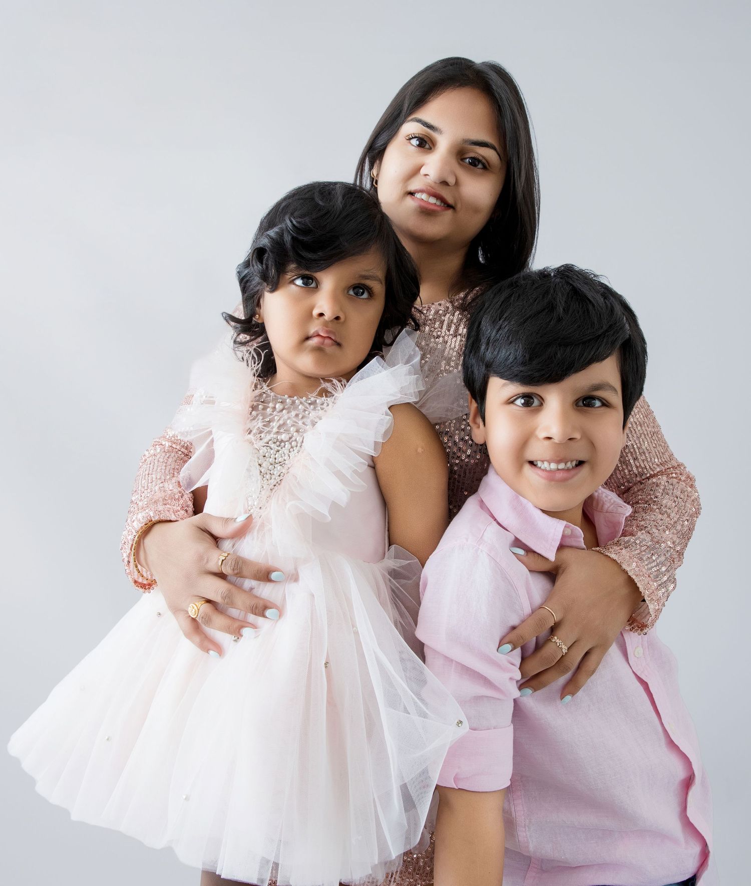 A mother poses with her two young children in pink outfits against a white background in a studio portrait.