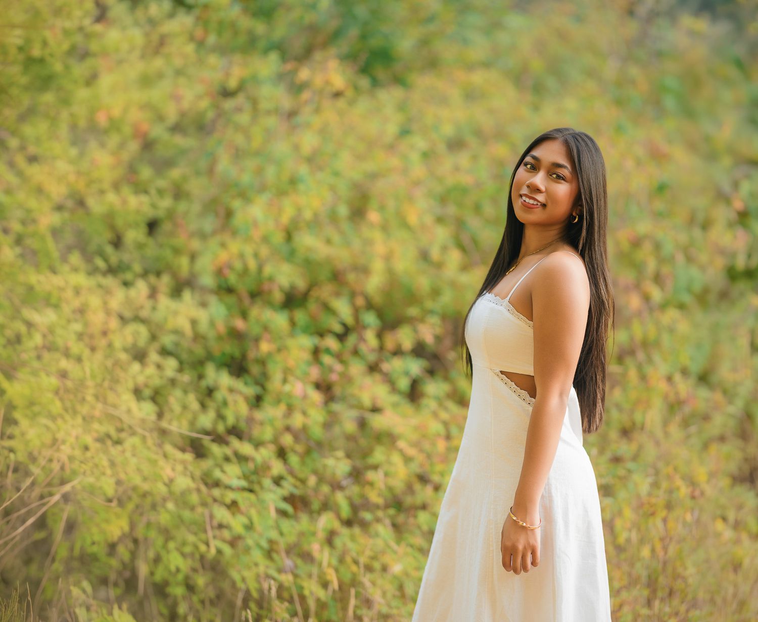 Young woman in white dress smiling outdoors with lush green and yellow foliage in background.