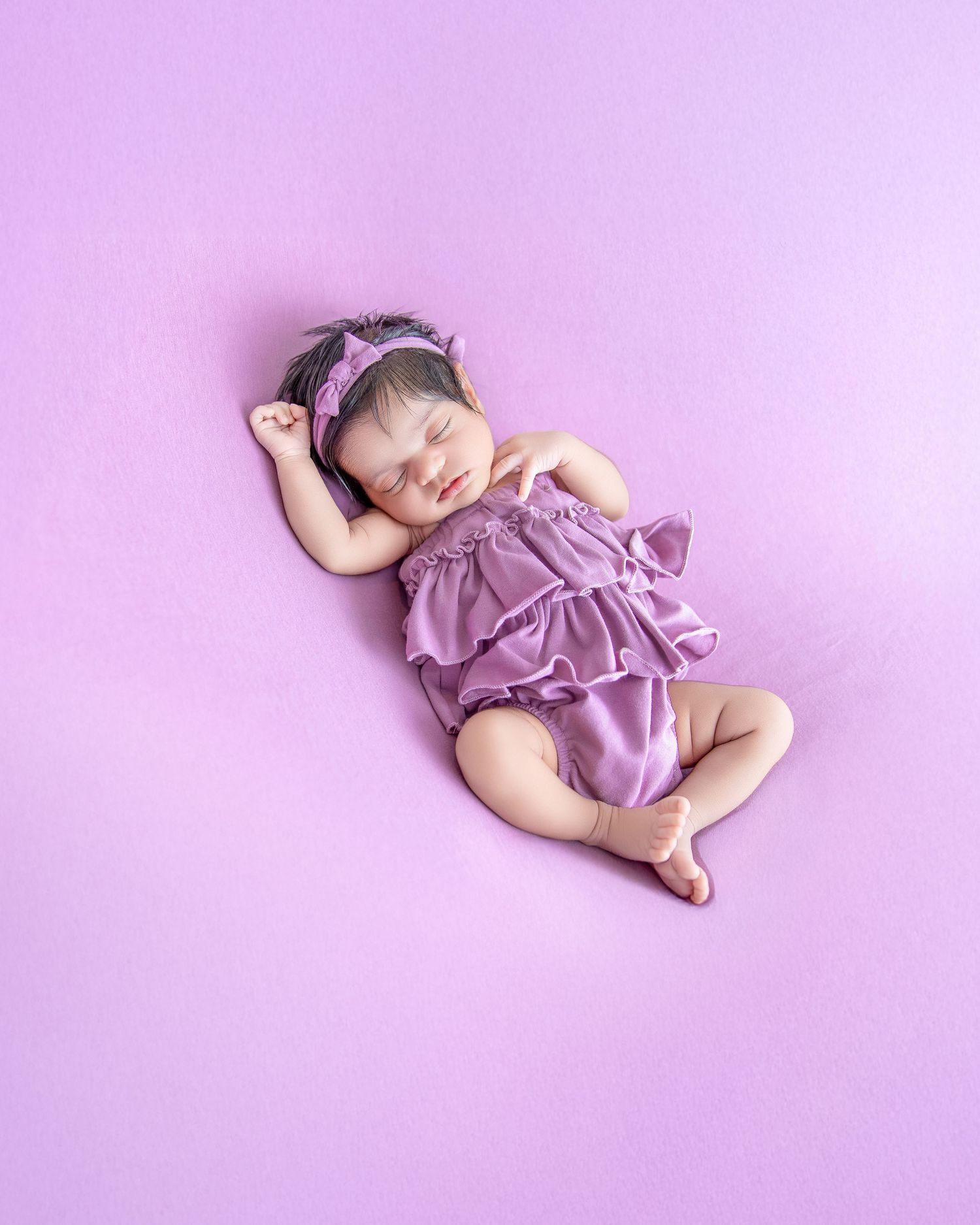 Sleeping newborn baby girl in purple ruffled romper with matching headband on lavender background.