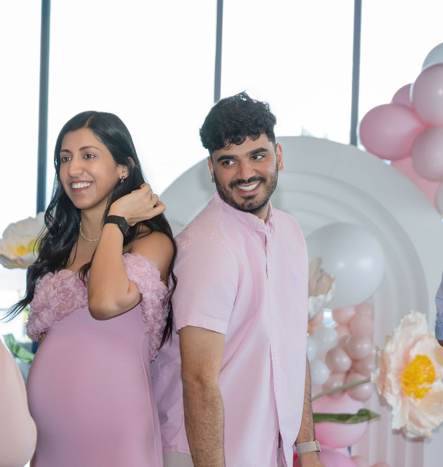 Smiling couple in pink outfits posing at a baby shower with balloon arch and floral decorations.