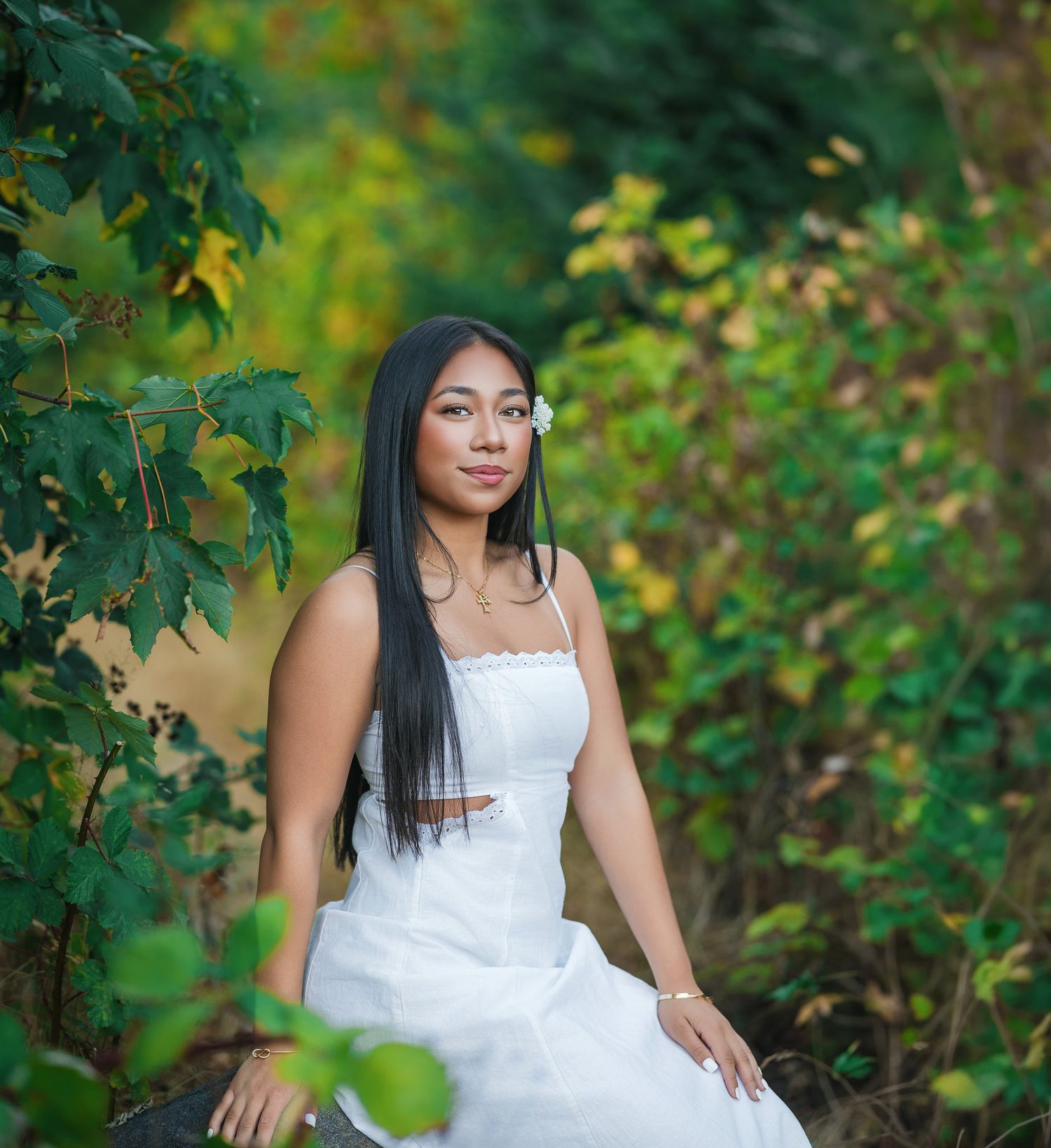 Young woman in white dress sits among lush green foliage with flower in hair.