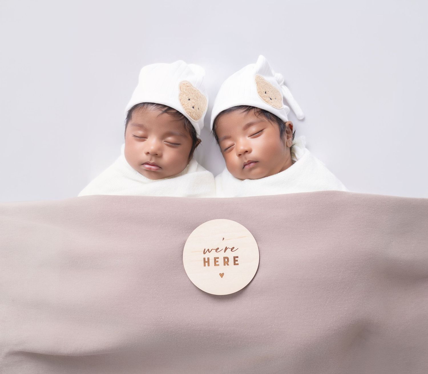 Two sleeping newborn twins wrapped in white swaddles wearing bear hats, lying together with a milestone disc reading we're here.