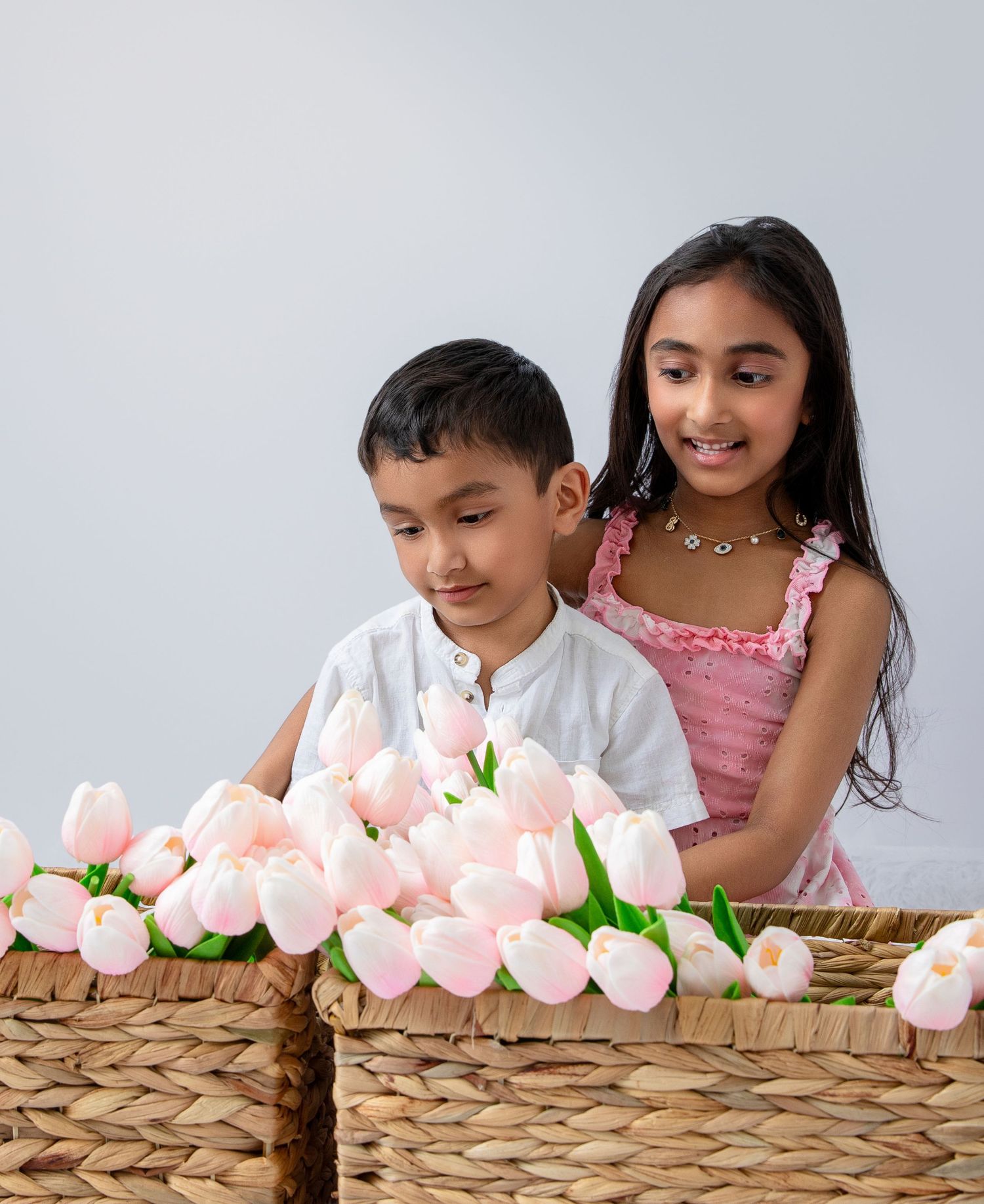 Two children admiring a basket of pink tulips, boy in white shirt and girl in pink dress against white background.