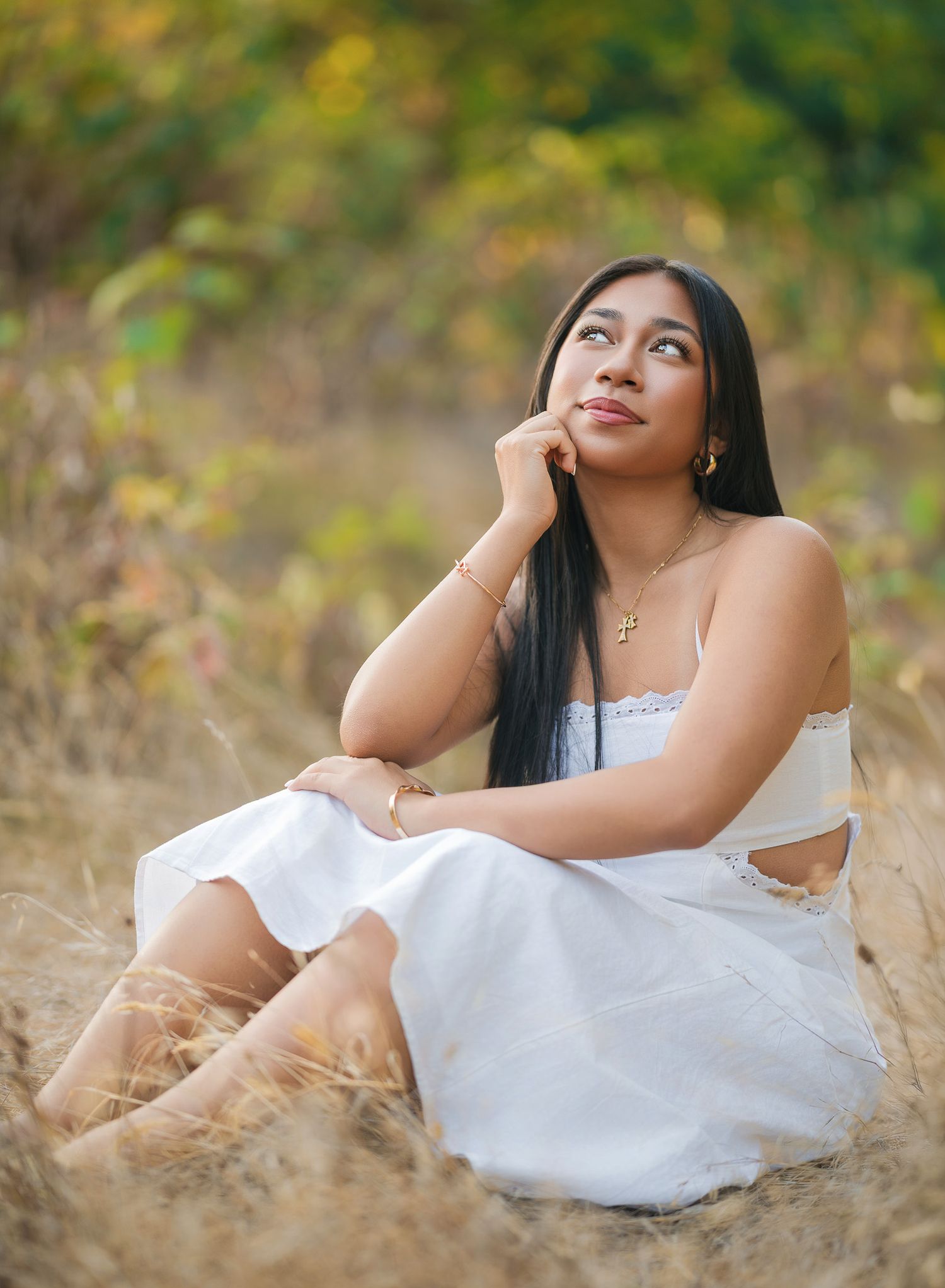 Young woman in white dress sitting in golden field, gazing upward thoughtfully, with long dark hair and gold jewelry.
