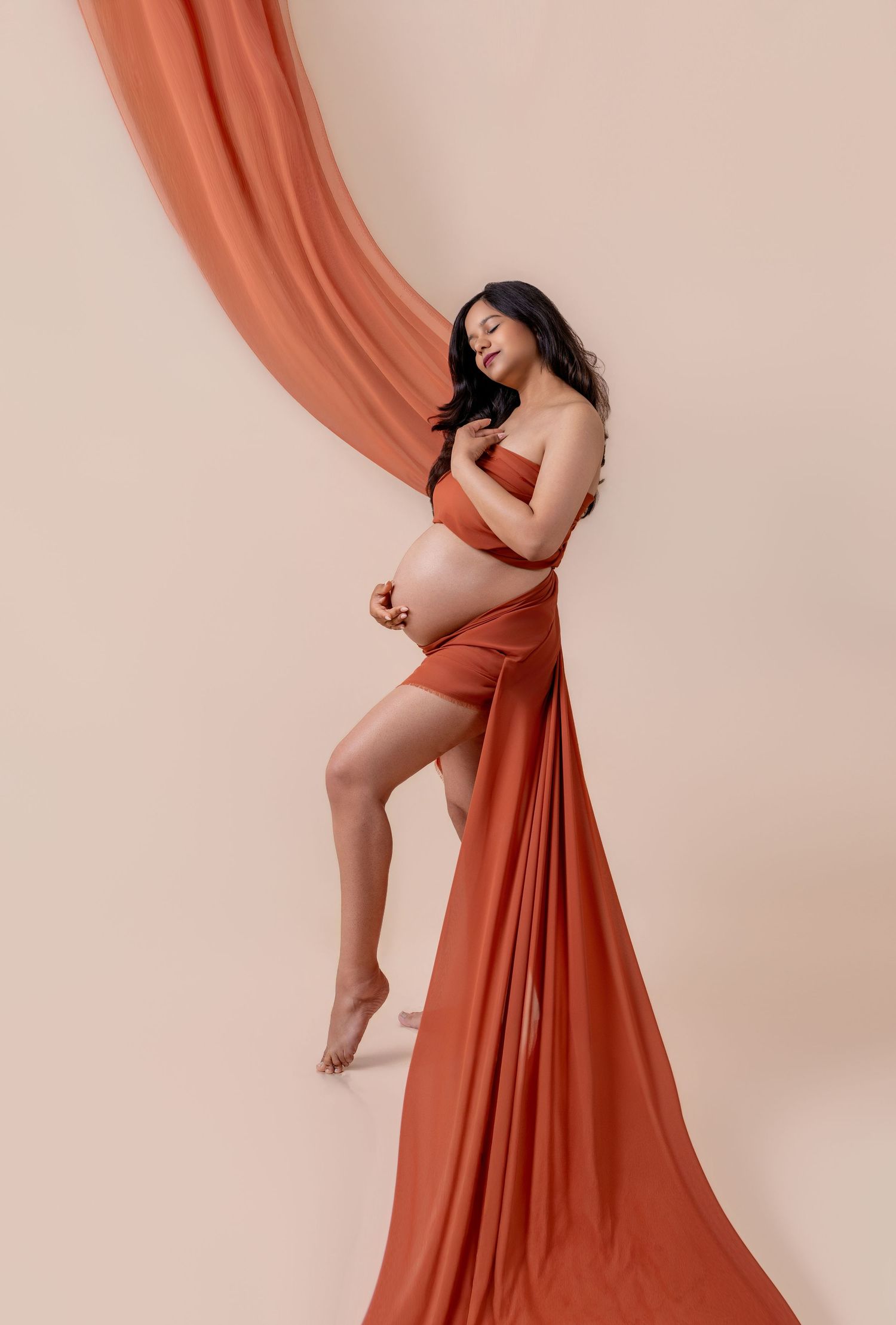 Pregnant woman in terracotta fabric wrap poses gracefully against a beige studio backdrop in a maternity photo shoot.