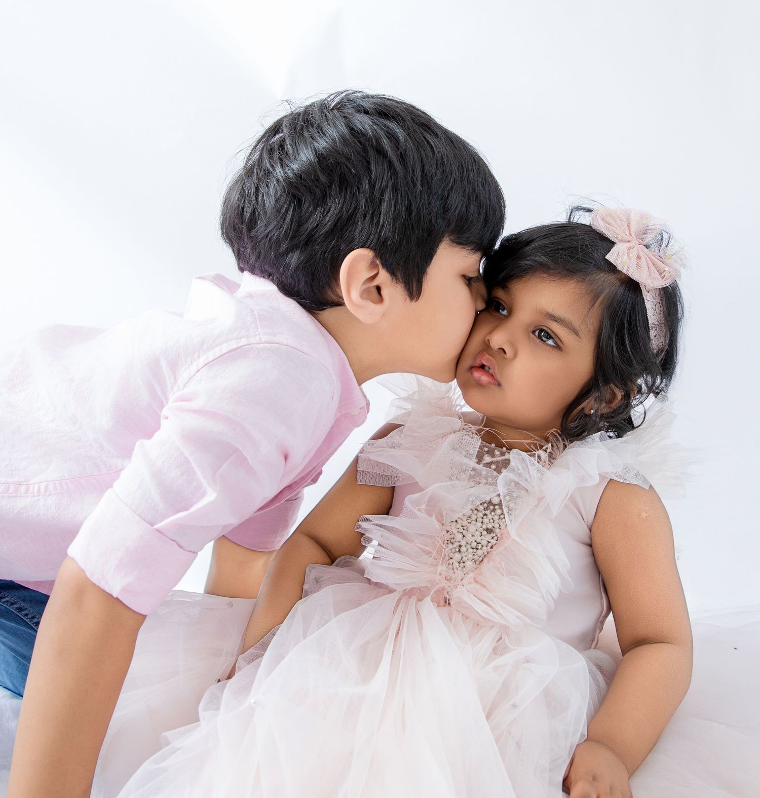 A young boy in pink shirt gently kisses the cheek of a toddler girl in a white tulle dress with pink bow.