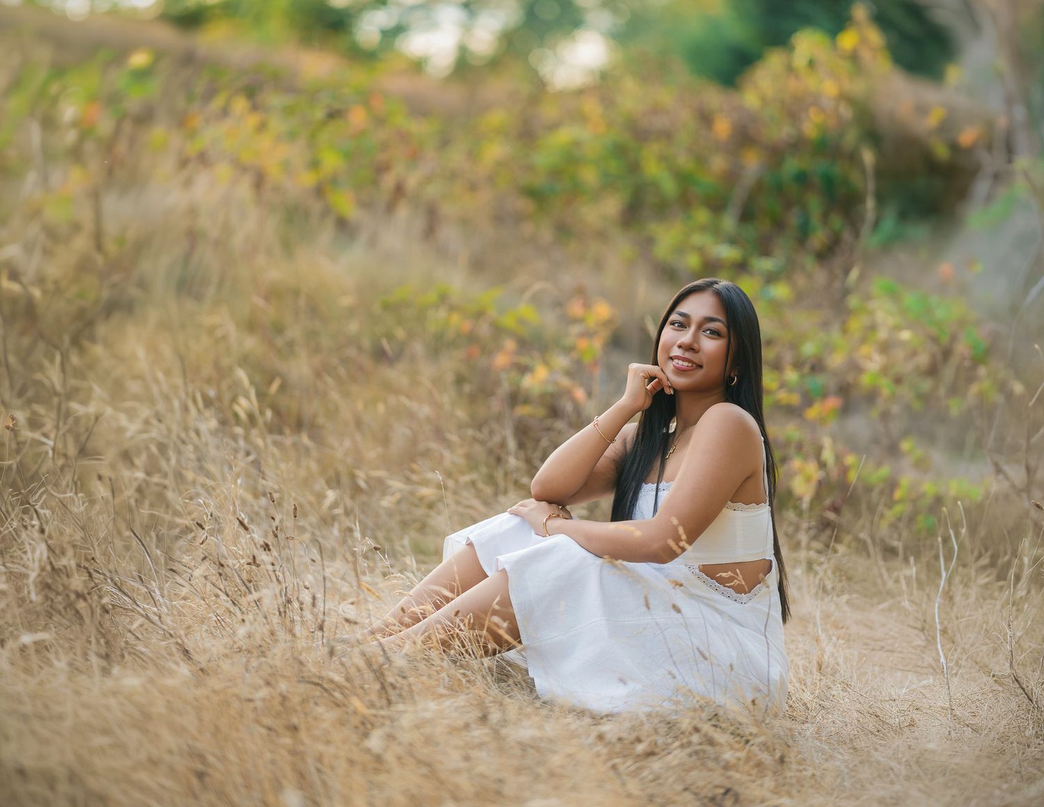Young woman in white dress sitting gracefully in golden dry grass field with autumn foliage background.