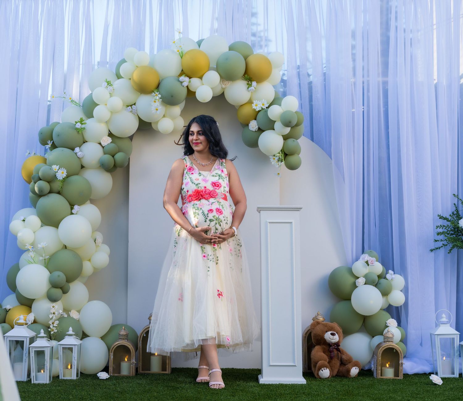 Woman in floral dress poses under balloon arch with green, white, and yellow balloons at elegant outdoor event.