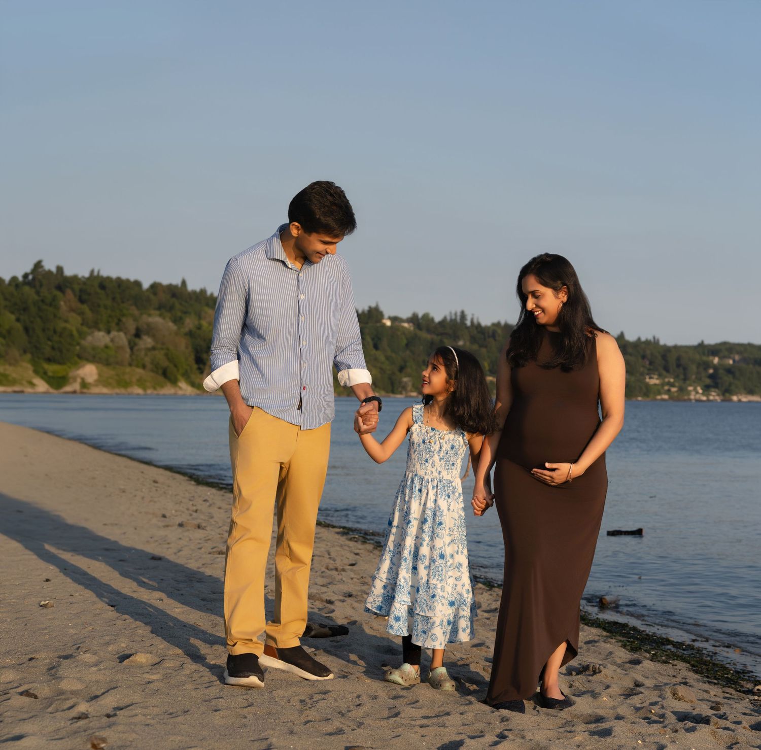 A family of three poses on a sandy beach at sunset, with a pregnant woman in a dark dress.