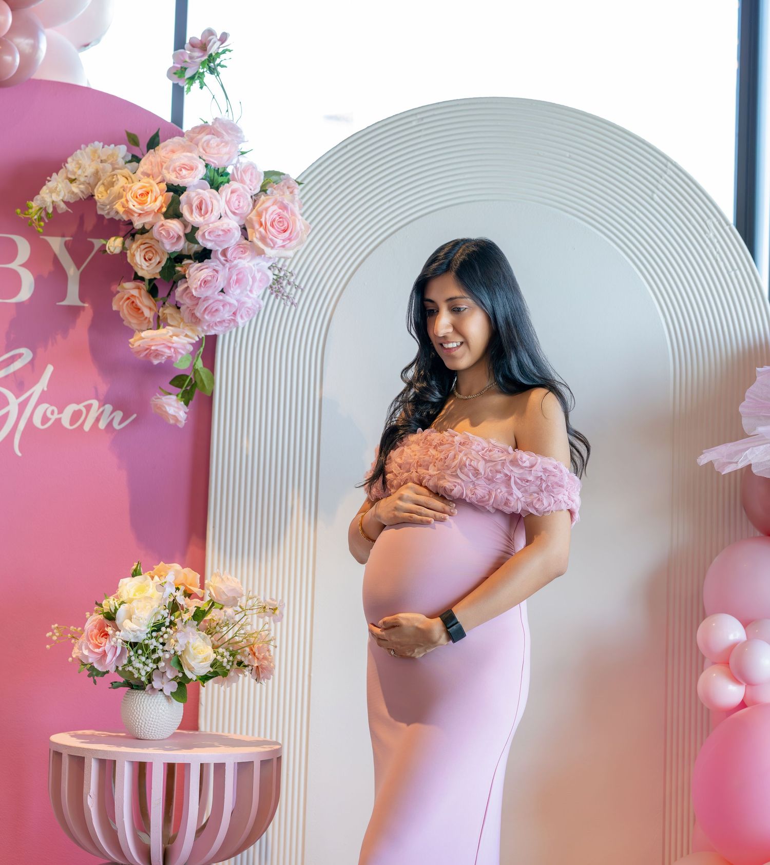 Pregnant woman in pink off-shoulder gown at baby shower with floral arrangements and pink balloon decorations.