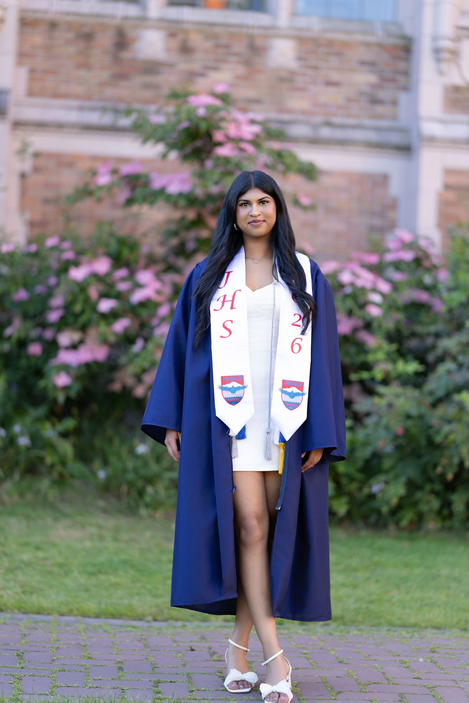 Young woman in navy blue graduation gown and white stole poses outdoors near blooming pink flowers and brick building.