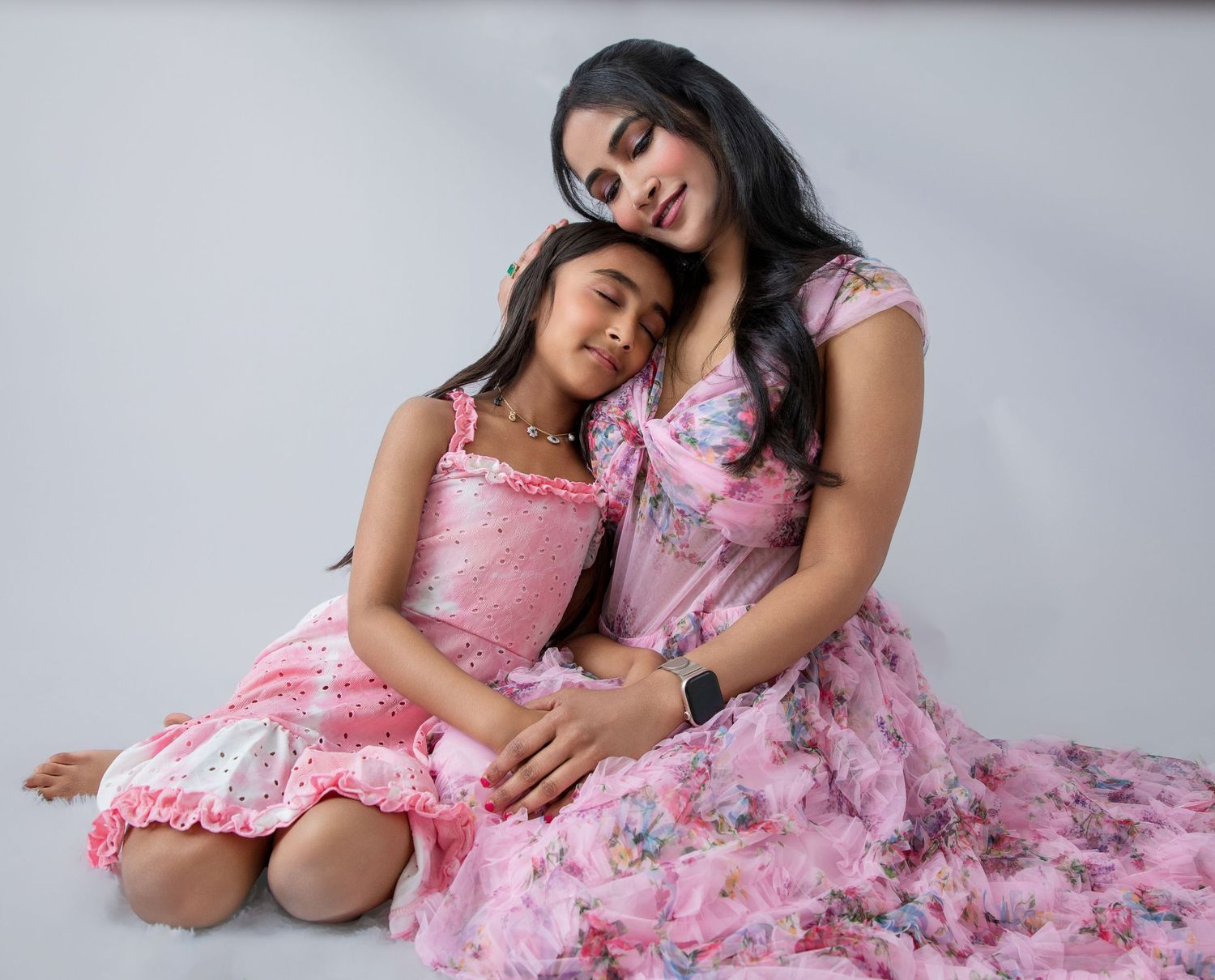 A woman and young girl in matching pink floral dresses pose together in a studio portrait.