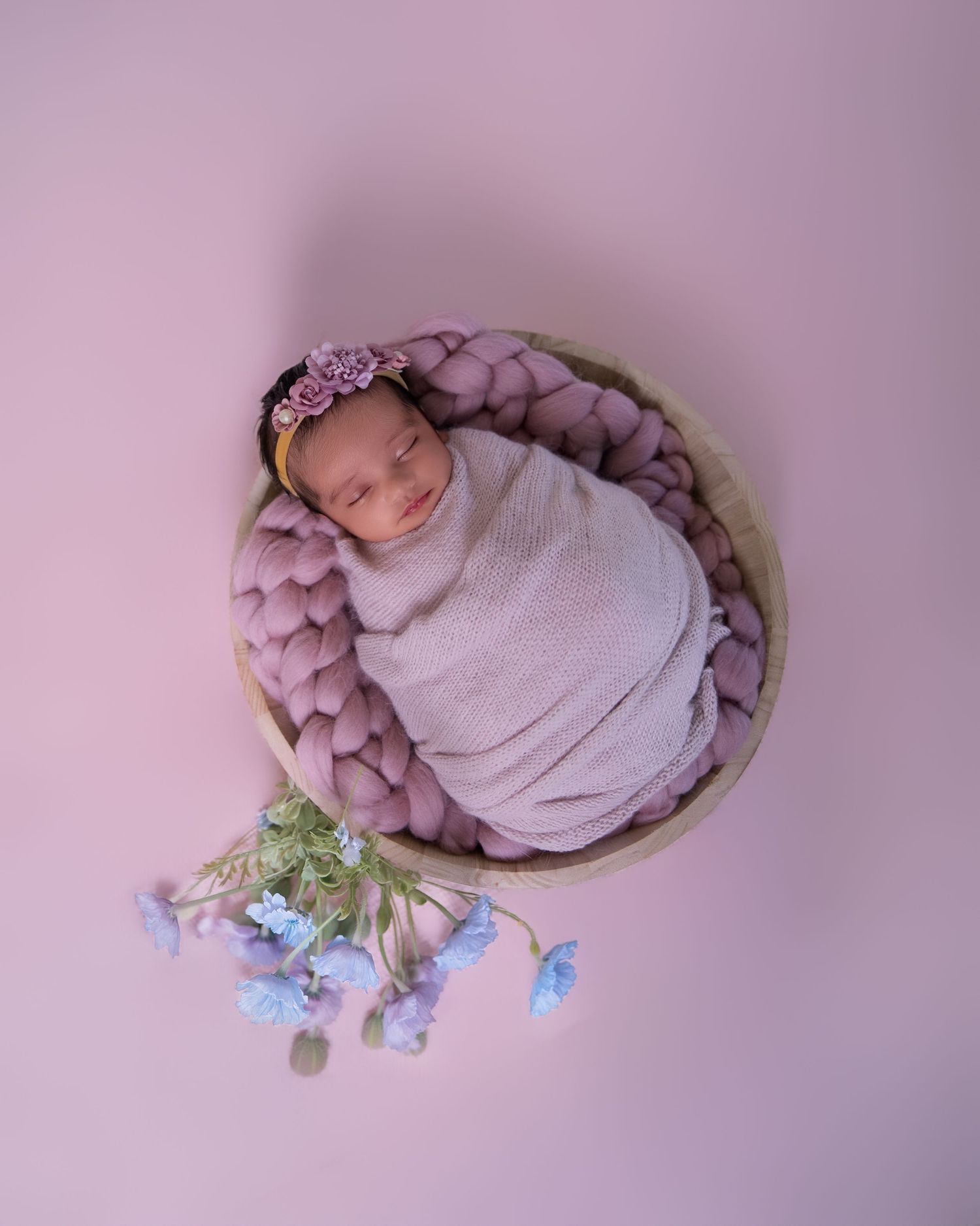 Newborn baby swaddled in lavender wrap sleeping in a round wooden bowl with chunky knit and blue flowers.