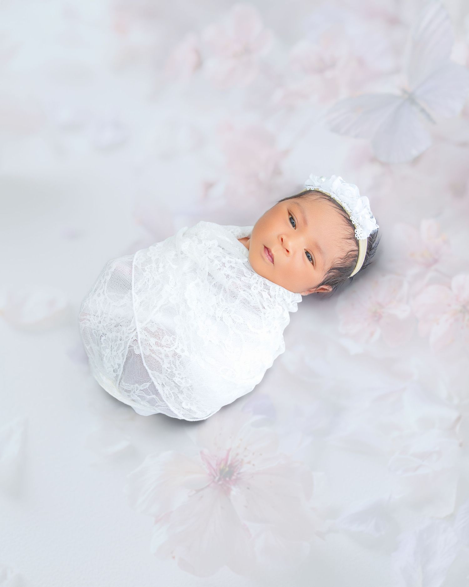 Peaceful newborn baby swaddled in white lace wrap with headband on soft floral backdrop.