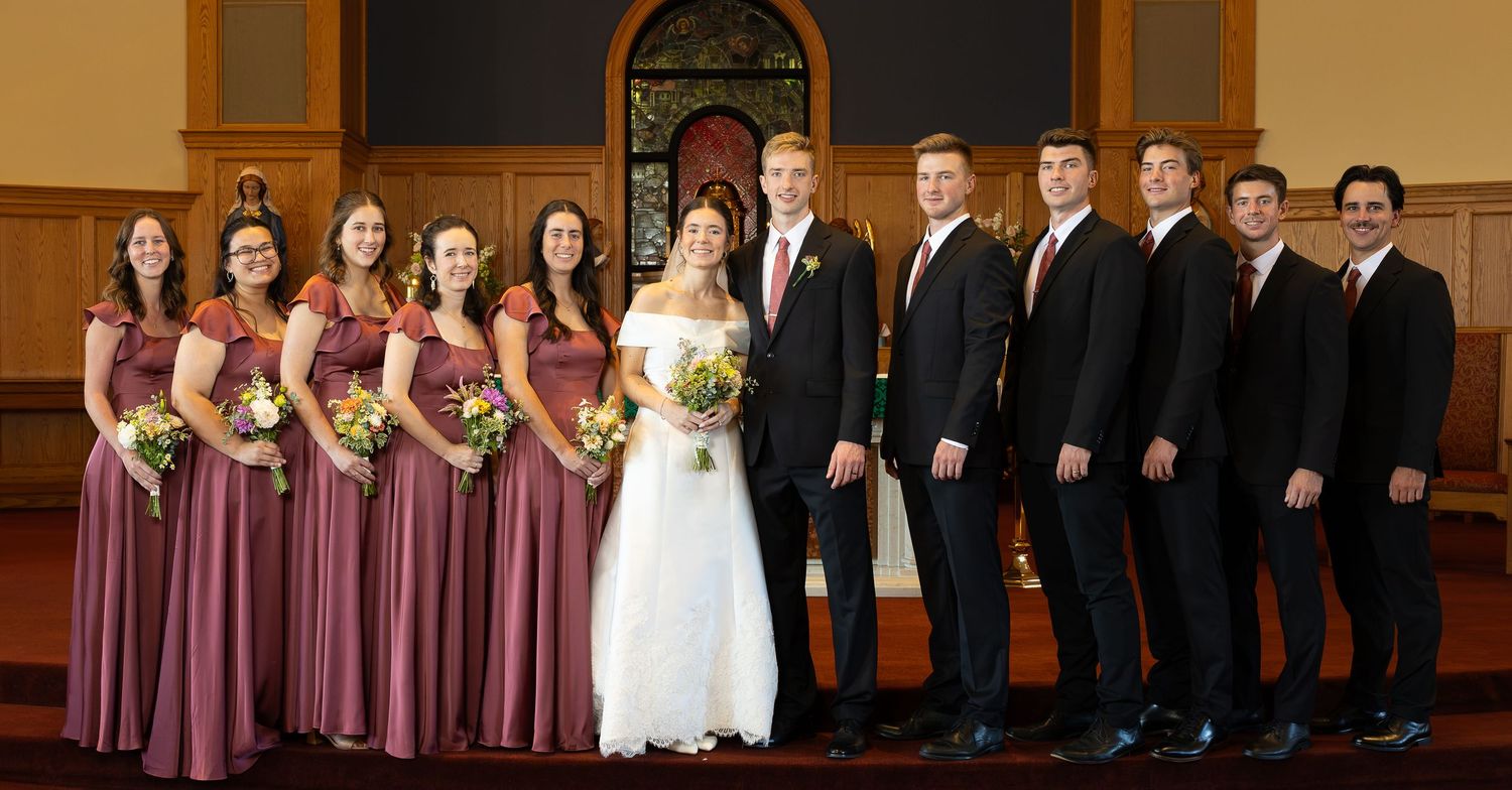 Wedding party posing in church with bridesmaids in burgundy gowns, groomsmen in black suits, and bride and groom in center.