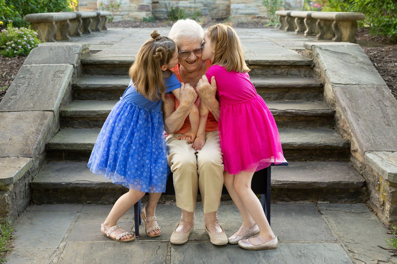 Elderly woman sitting on stone steps receiving kisses from two young girls in blue and pink dresses outdoors.