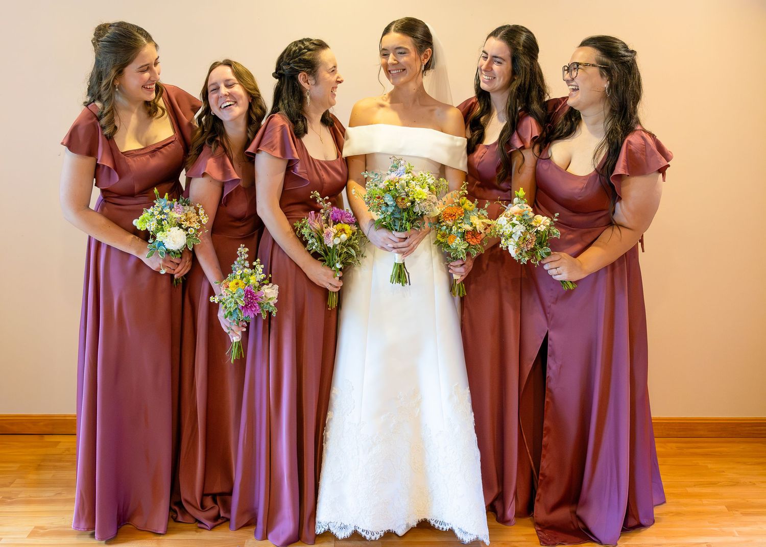 Bride in white lace gown smiles with four bridesmaids in dusty rose dresses holding colorful bouquets.